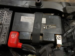 Close-up of a motorcycle battery secured with a strap, red terminal visible