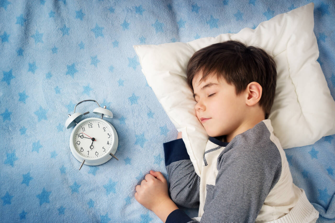 A young boy sleeping peacefully on a white pillow next to a white alarm clock on a blue star-patterned blanket.
