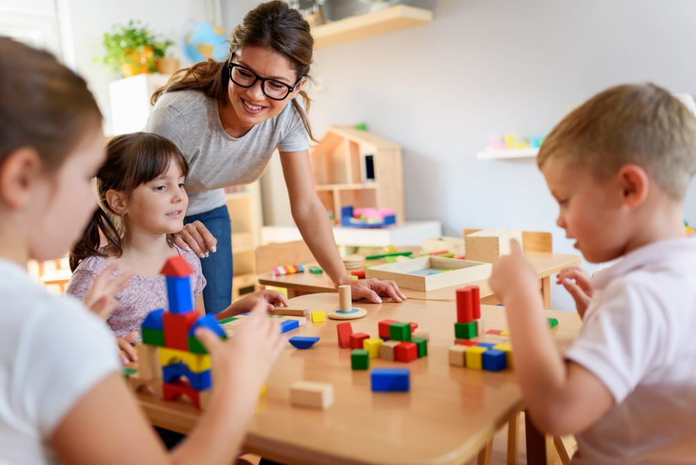From Interest to Enrollment: Best Practices for Marketing Your Family Child Care Business Smiling teacher in glasses watching three young children build structures with colorful wooden blocks in a classroom.