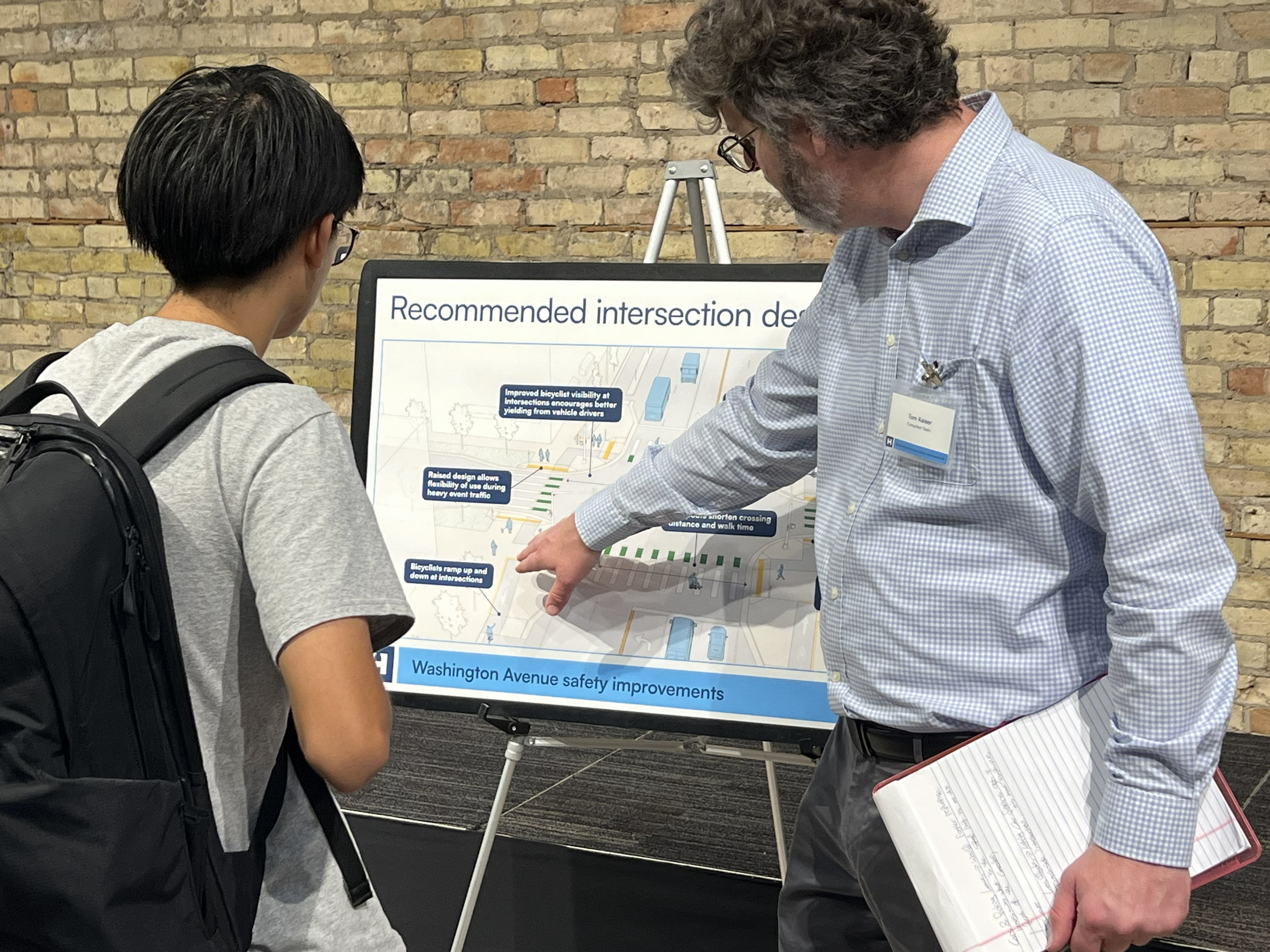 A person with a backpack is looking at a poster. A project staff member is pointing at the poster.