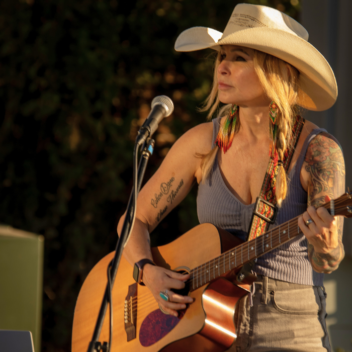 A woman wearing a wide-brimmed hat holds an acoustic guitar while singing into a microphone. She has colorful earrings and visible tattoos on her arms.