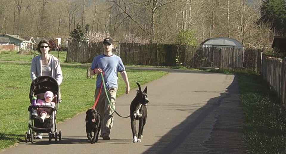 Dos personas caminan con un cochecito de bebé y unos perros por un sendero pavimentado que atraviesa una zona de césped, alejado de la carretera, como ejemplo de un sendero fuera del corredor.
