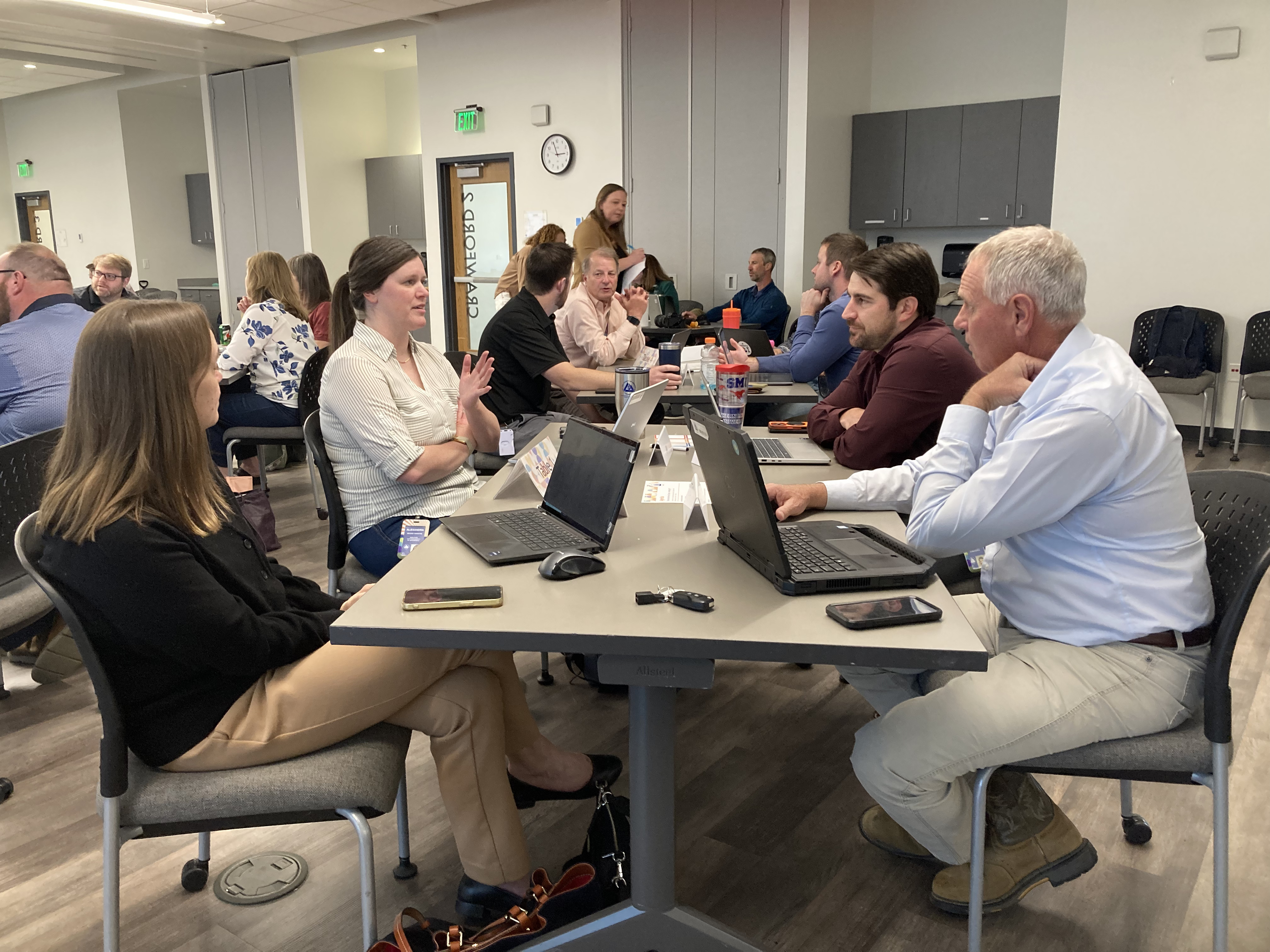 A group of four people sit around a table deep in discussion. Laptops are open to capture notes.