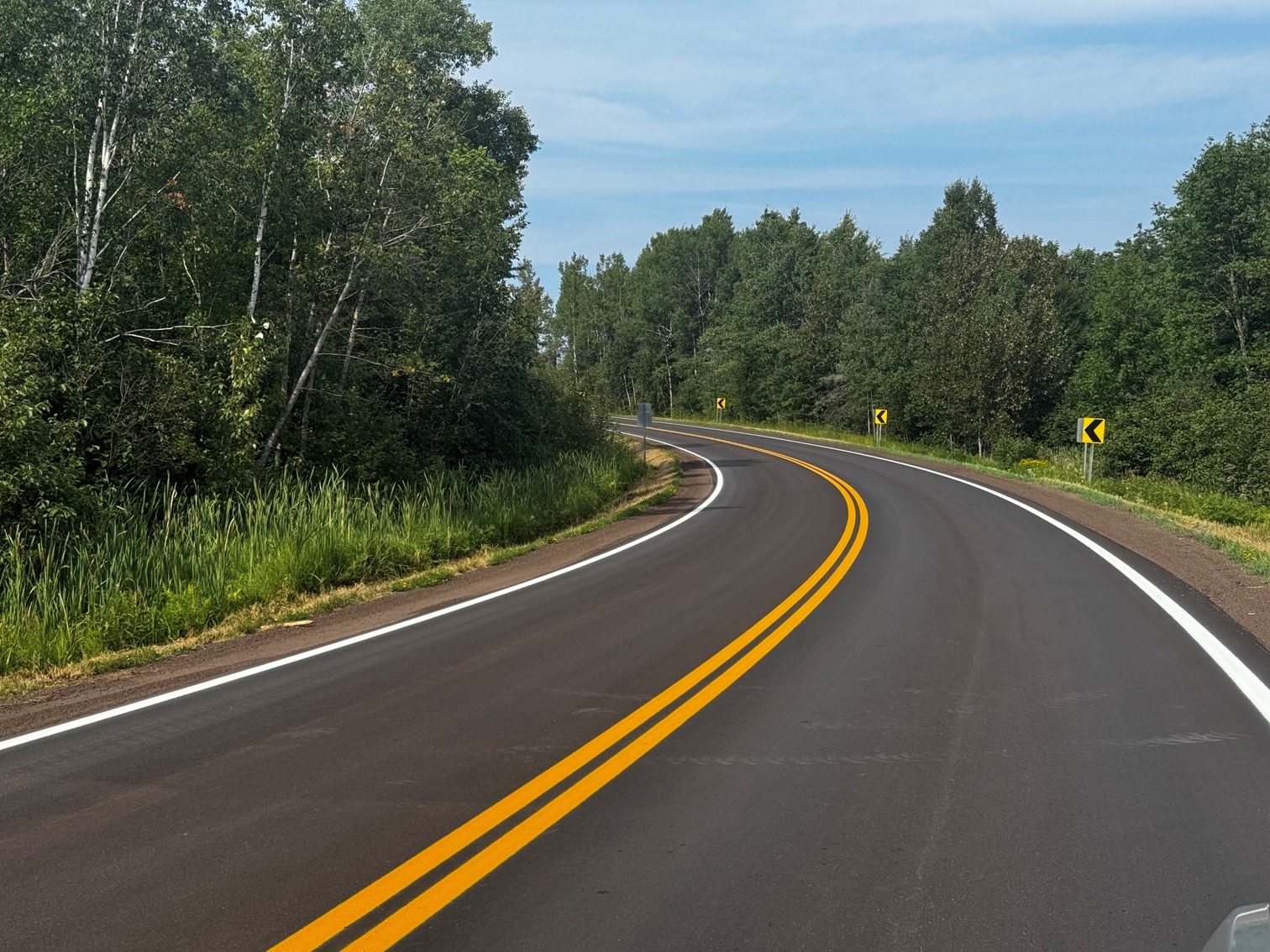 A winding asphalt road with double yellow lines, bordered by lush greenery and trees under a partly cloudy sky.