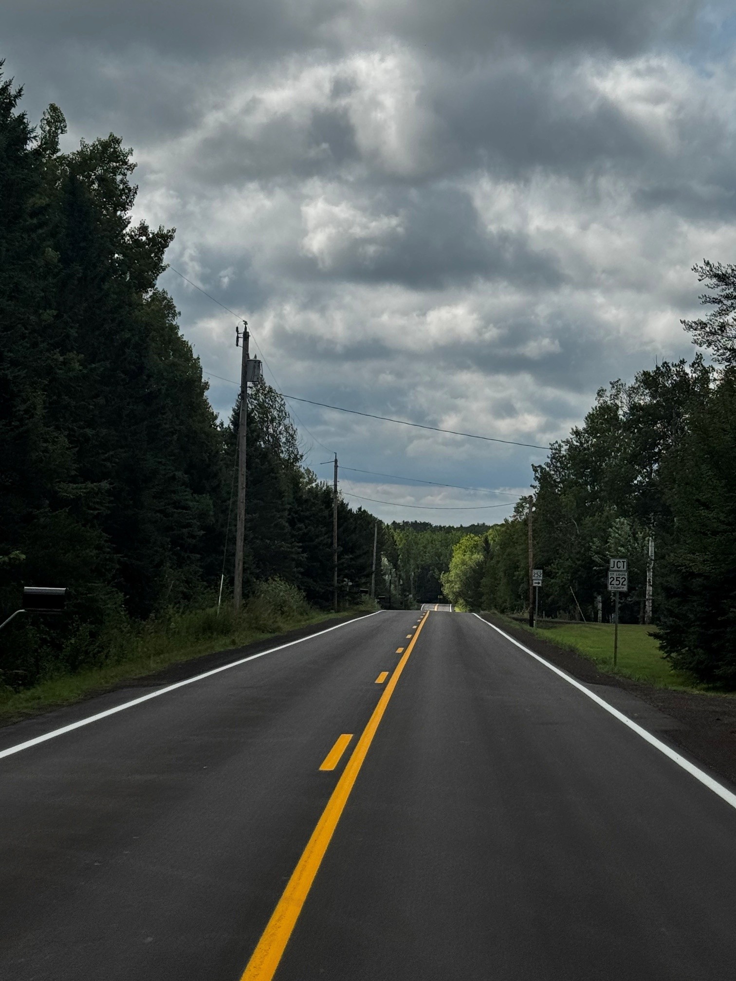 A freshly paved two-lane road winds through dense forest beneath dark, sunlit clouds, with utility poles and a junction sign on the right.
