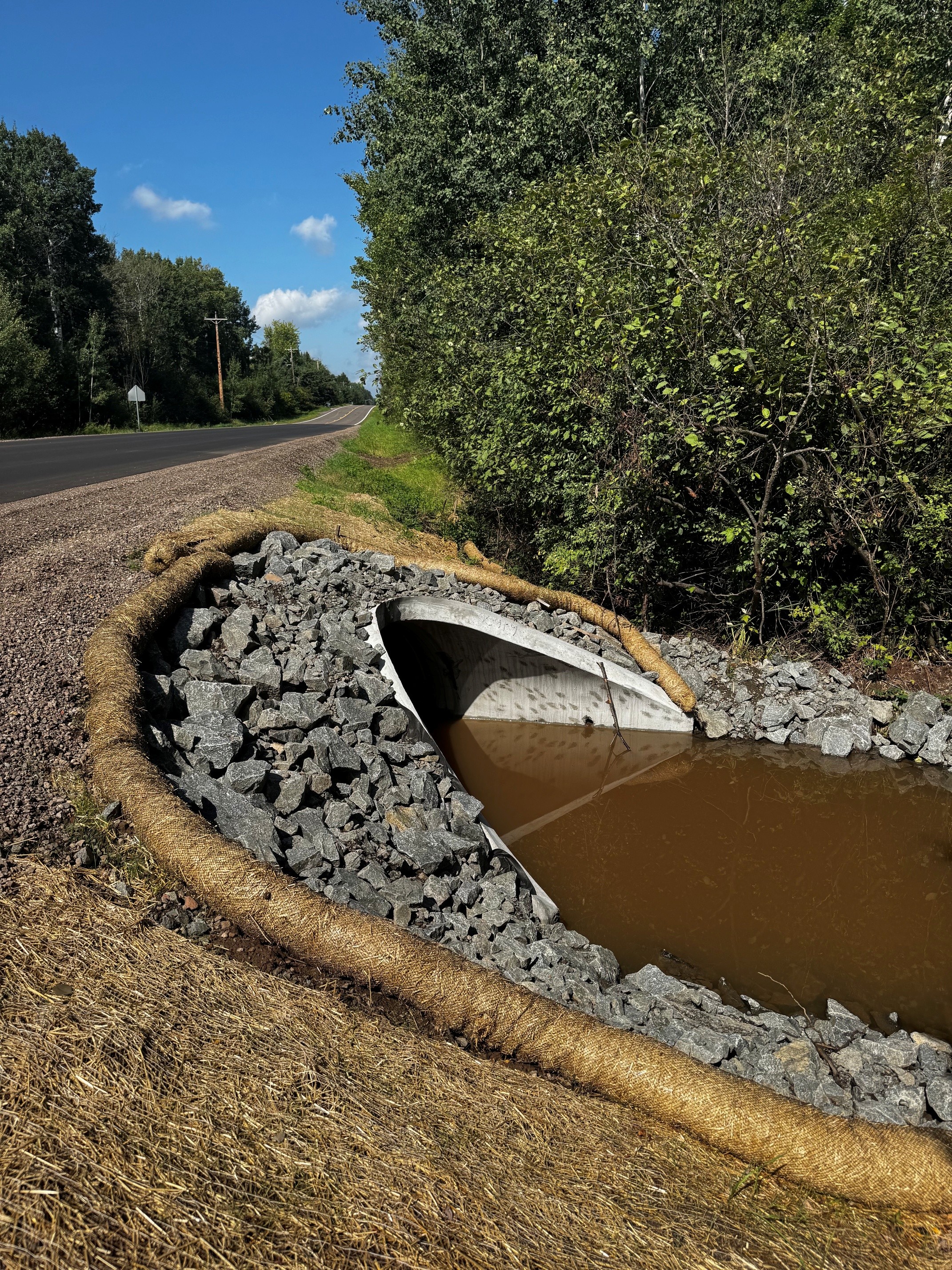 Concrete roadside culvert surrounded by gray rocks and straw erosion-control matting, with muddy water in the foreground and a wooded road beyond.