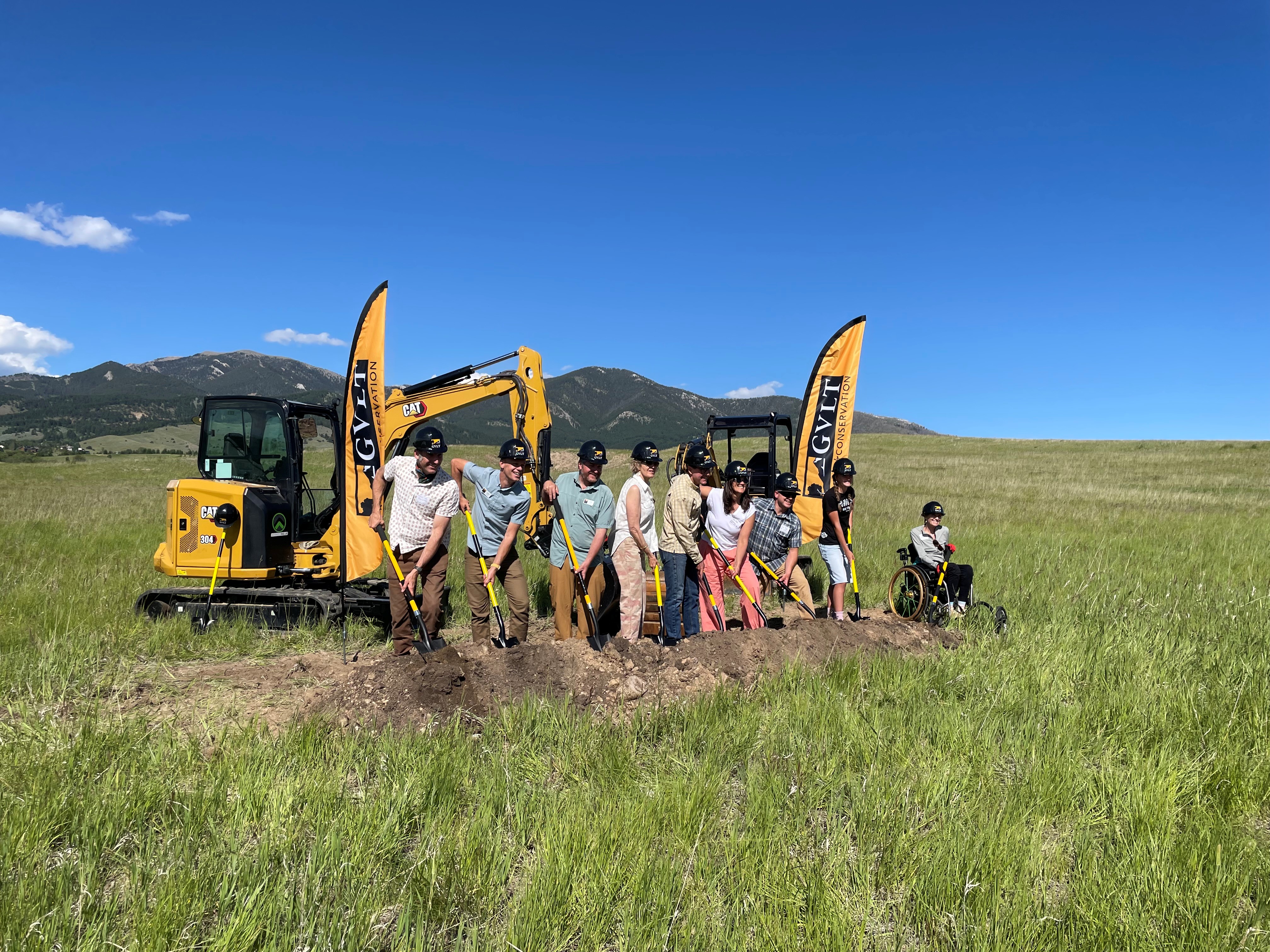 A group of people wearing hard hats shovel dirt with construction equipment in the background.