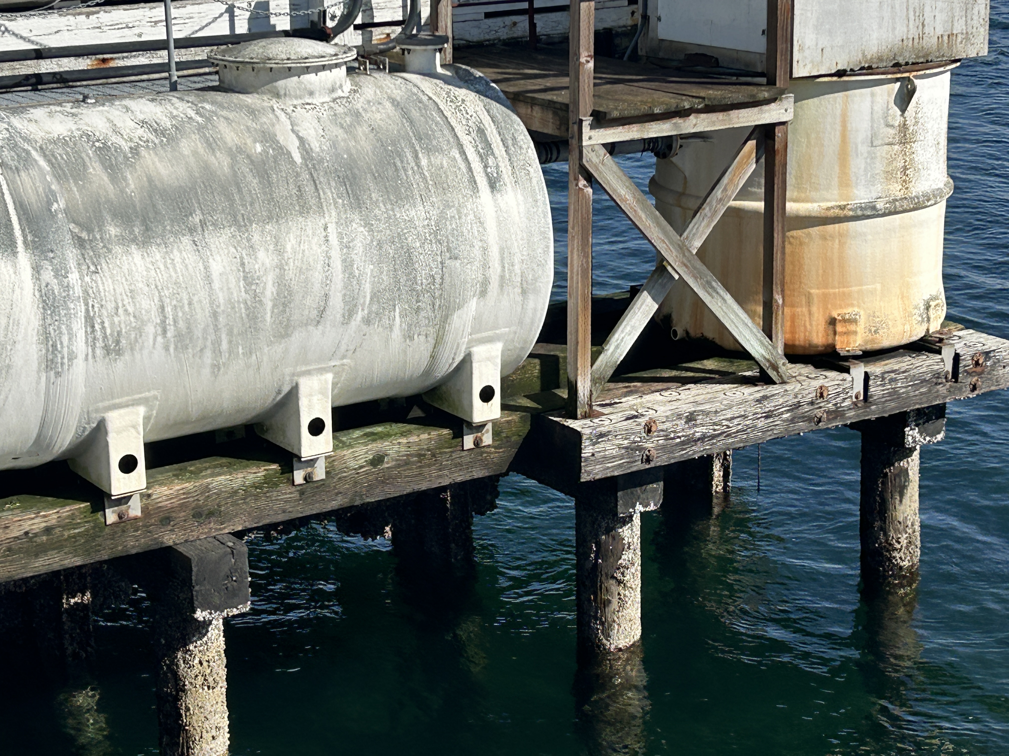A close-up view of a storage tank and metal infrastructure on top of aging piles with visible rust and algae or moss.