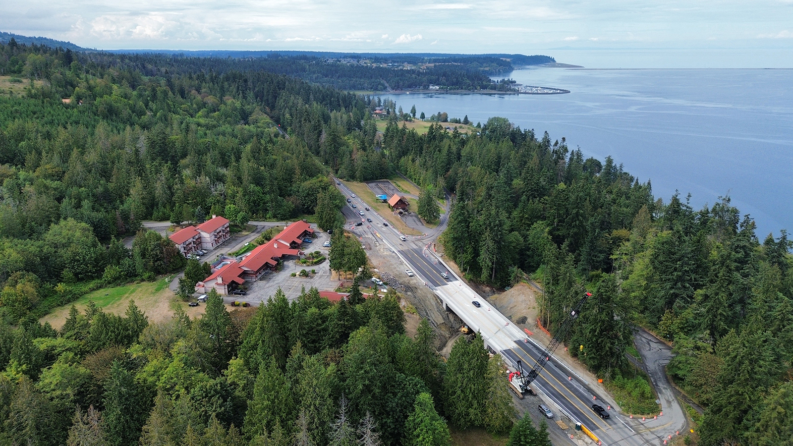 An aerial view of US 101 and the new Discovery Creek Bridge surrounded by conifer forests with Sequim Bay as a backdrop. WSDOT built a new bridge to improve fish passage and wildlife connectivity in Sequim. The vantage point looks northeast and includes Sequim Bay Lodge to the left of the road and the Olympic Discovery Trail to the right.