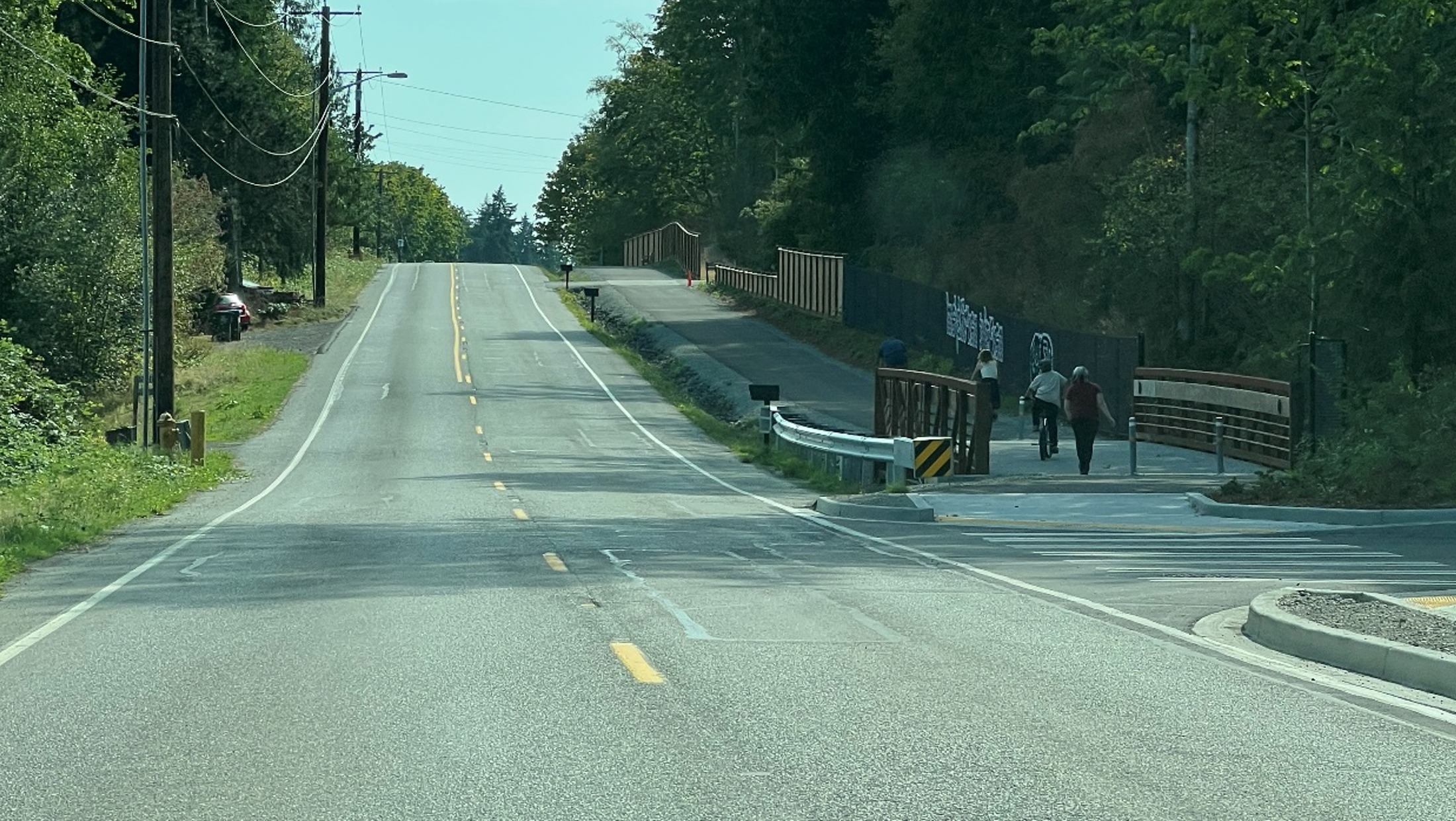 Two people walk on a paved path beside a road, separated from vehicle lanes by a roadside barrier.