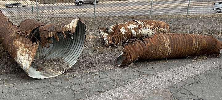 Large, rusty, broken stormwater pipes lie on dry ground near a cracked sidewalk and a chain-link fence.