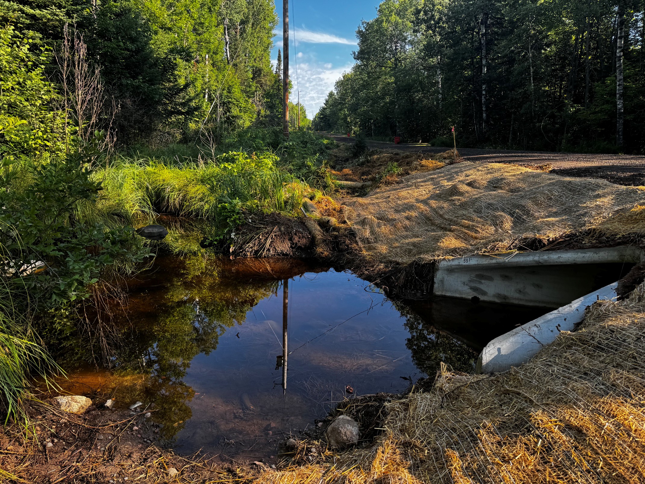 A shallow stream bordered by green vegetation, with a concrete culvert visible and a telephone pole in the background. Straw matting covers the ground near the water.