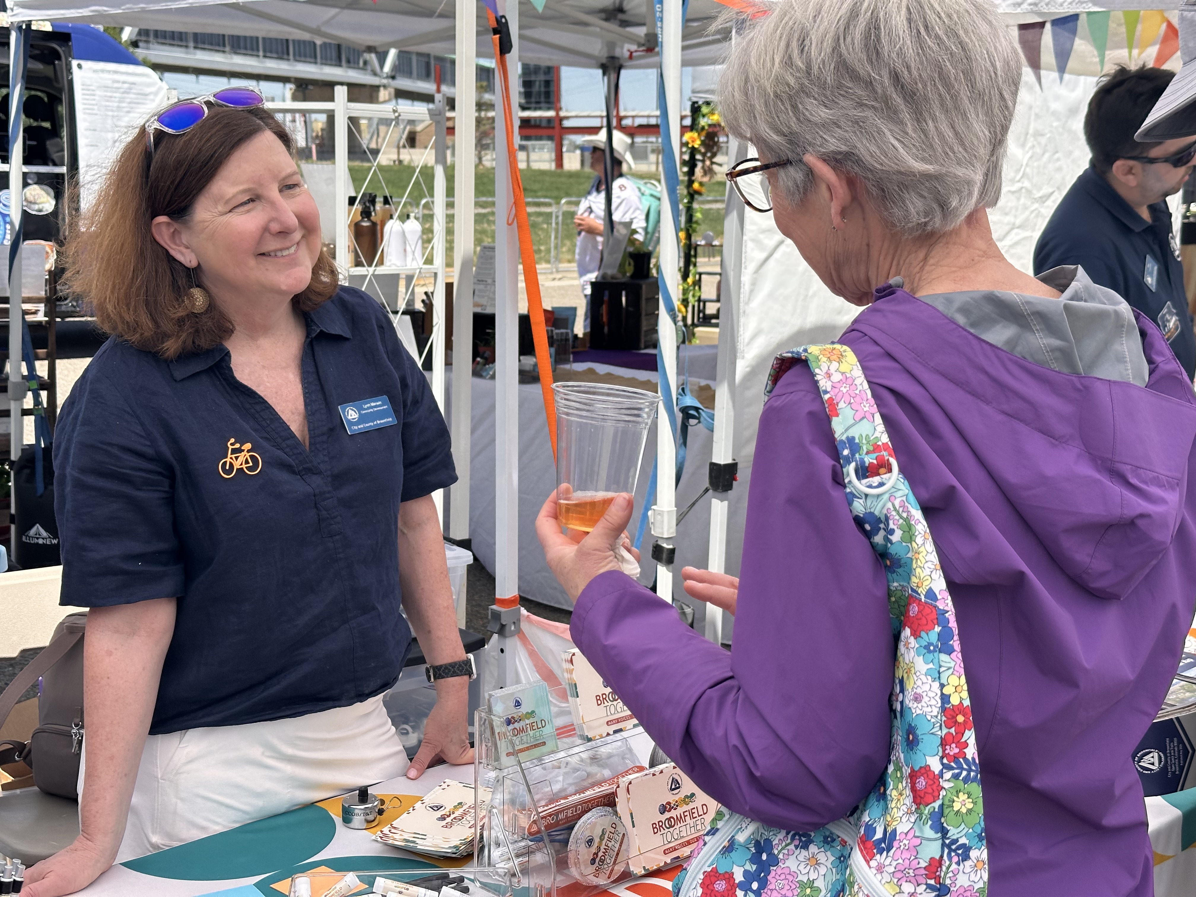A woman wearing a City and County of Broomfield name tag and a bicycle pin smiles while interacting with a visitor at an outdoor event booth. The table in front of her displays "Broomfield Together" brochures and promotional materials.