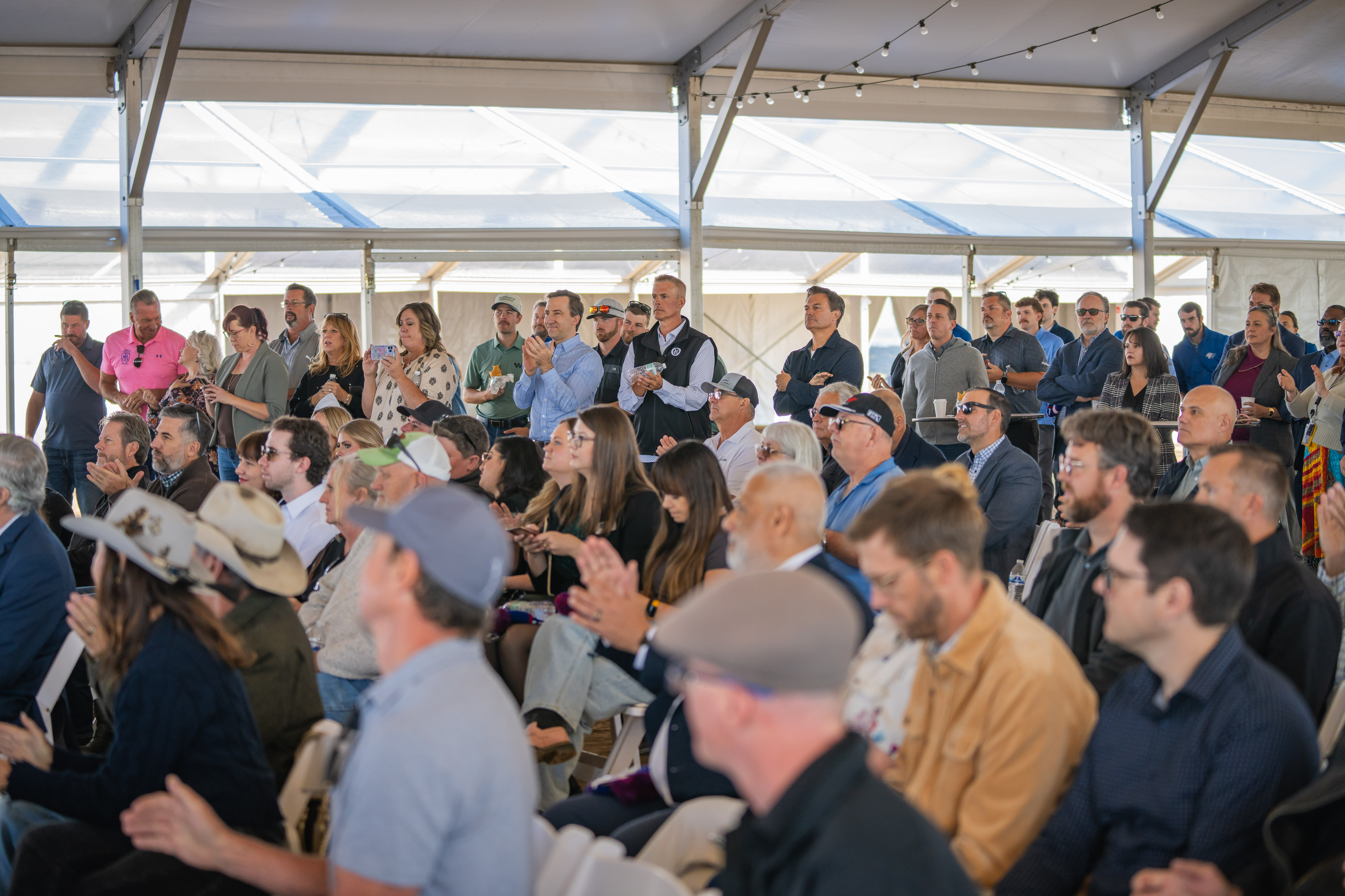 Audience of community members, city leaders, and partners seated and standing under a large tent, applauding during the West Greeley Catalyst Groundbreaking ceremony.
