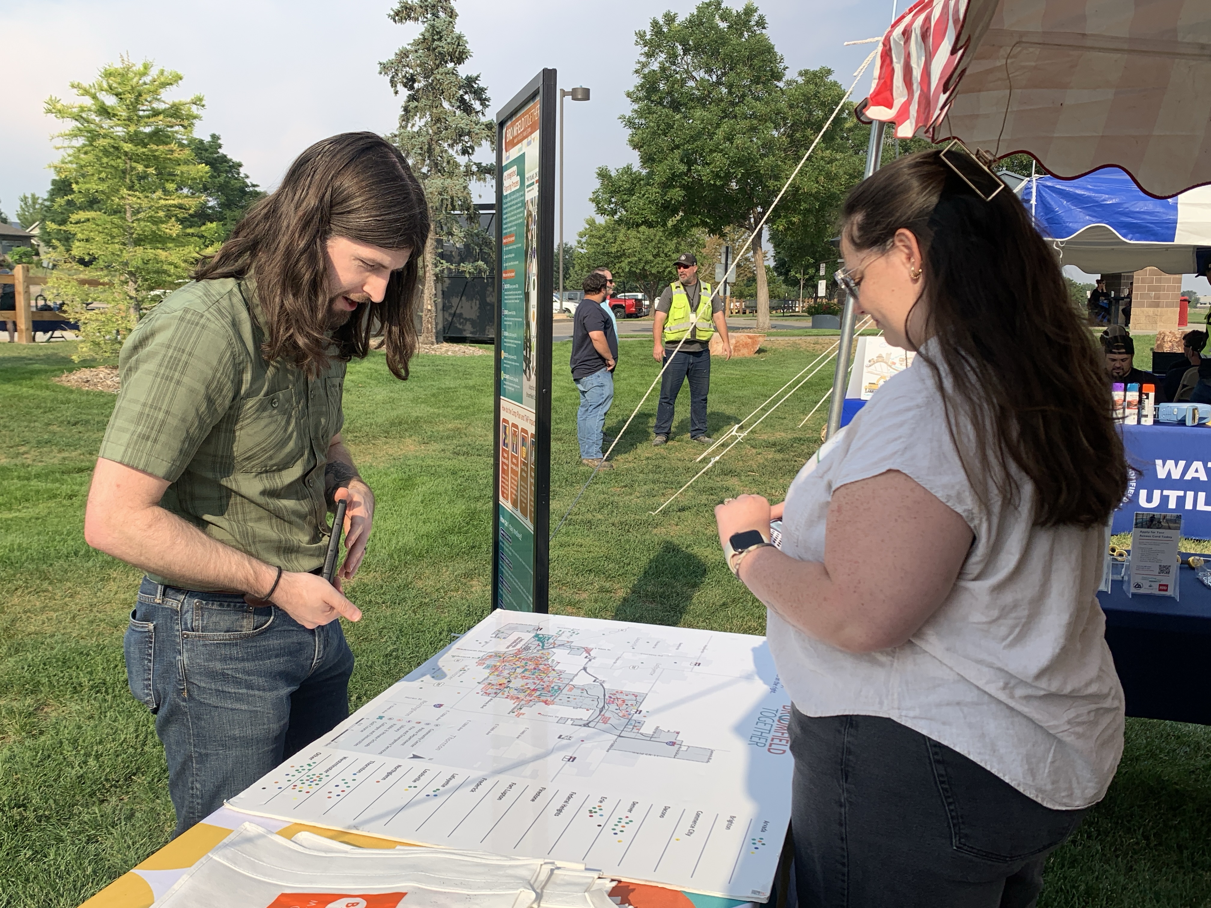 A man and a woman look at a large map titled "Broomfield Together" on a table at an outdoor community event. The man leans over the map while the woman stands opposite him. In the background, there is a tall informational sign and other people on a grassy field.