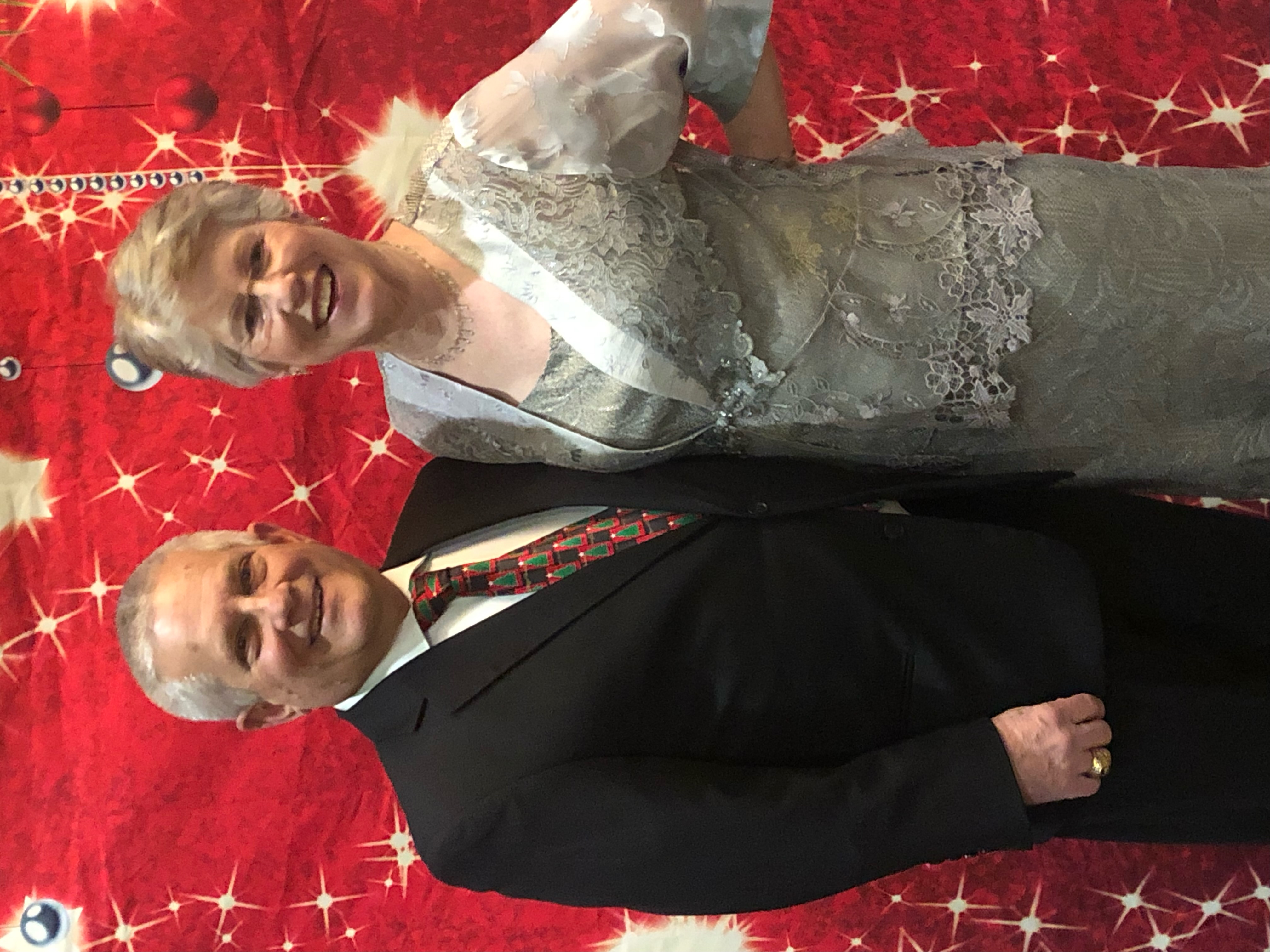 An older gentleman in a black suit and christmas tree tie stands smiling with his wife. She's in a festive silver dress. There's a red holiday back drop behind them.