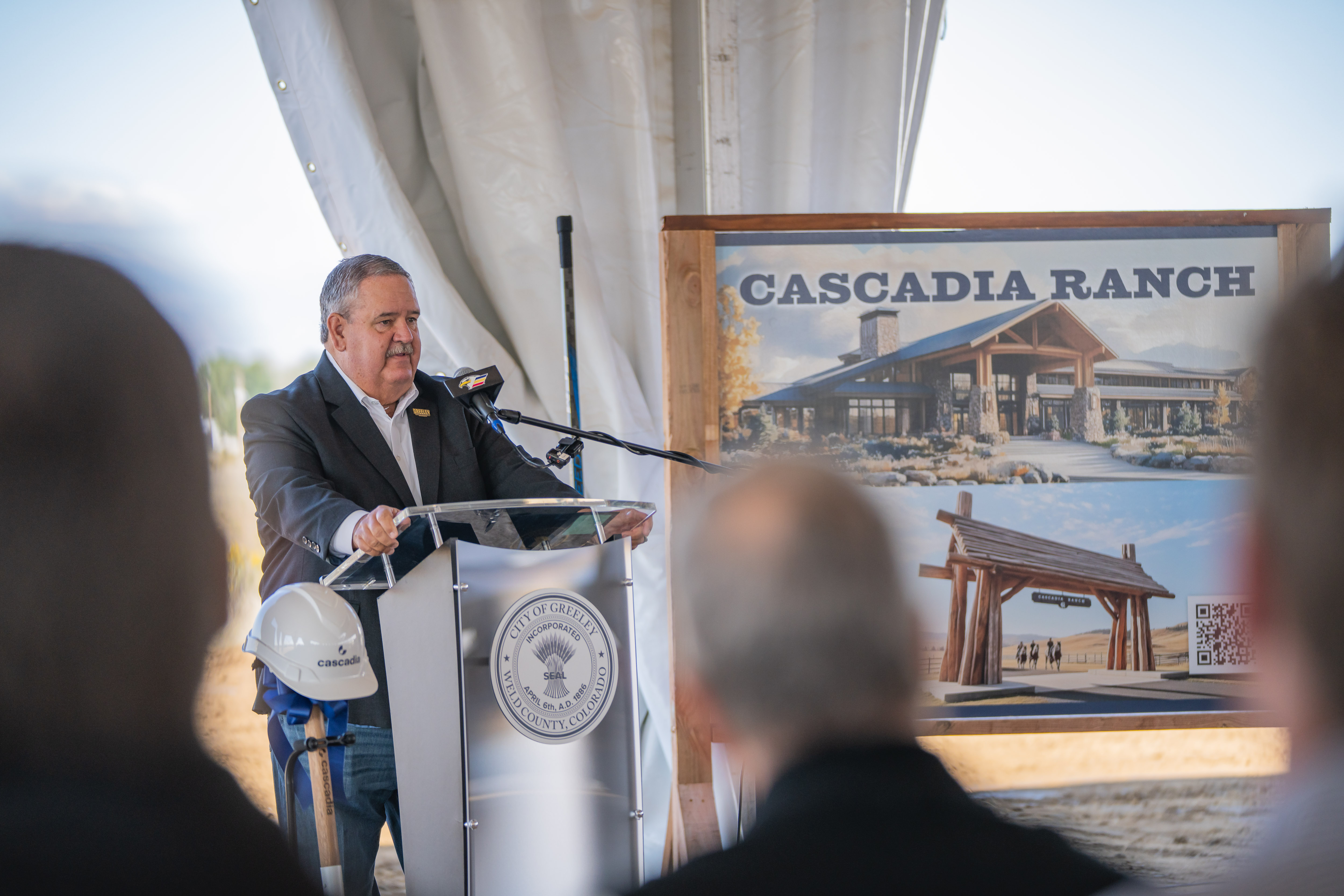 Mayor John Gates delivers speech to spectators. A sign for the Cascadia ranch stands behind him.