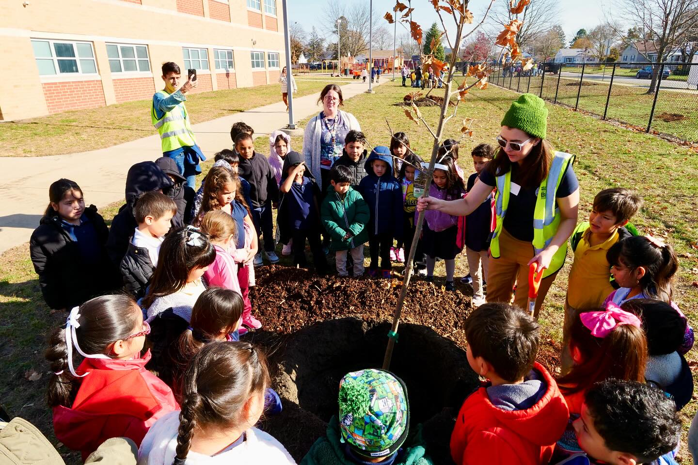 A person planting a tree while school children gather around to participate.
