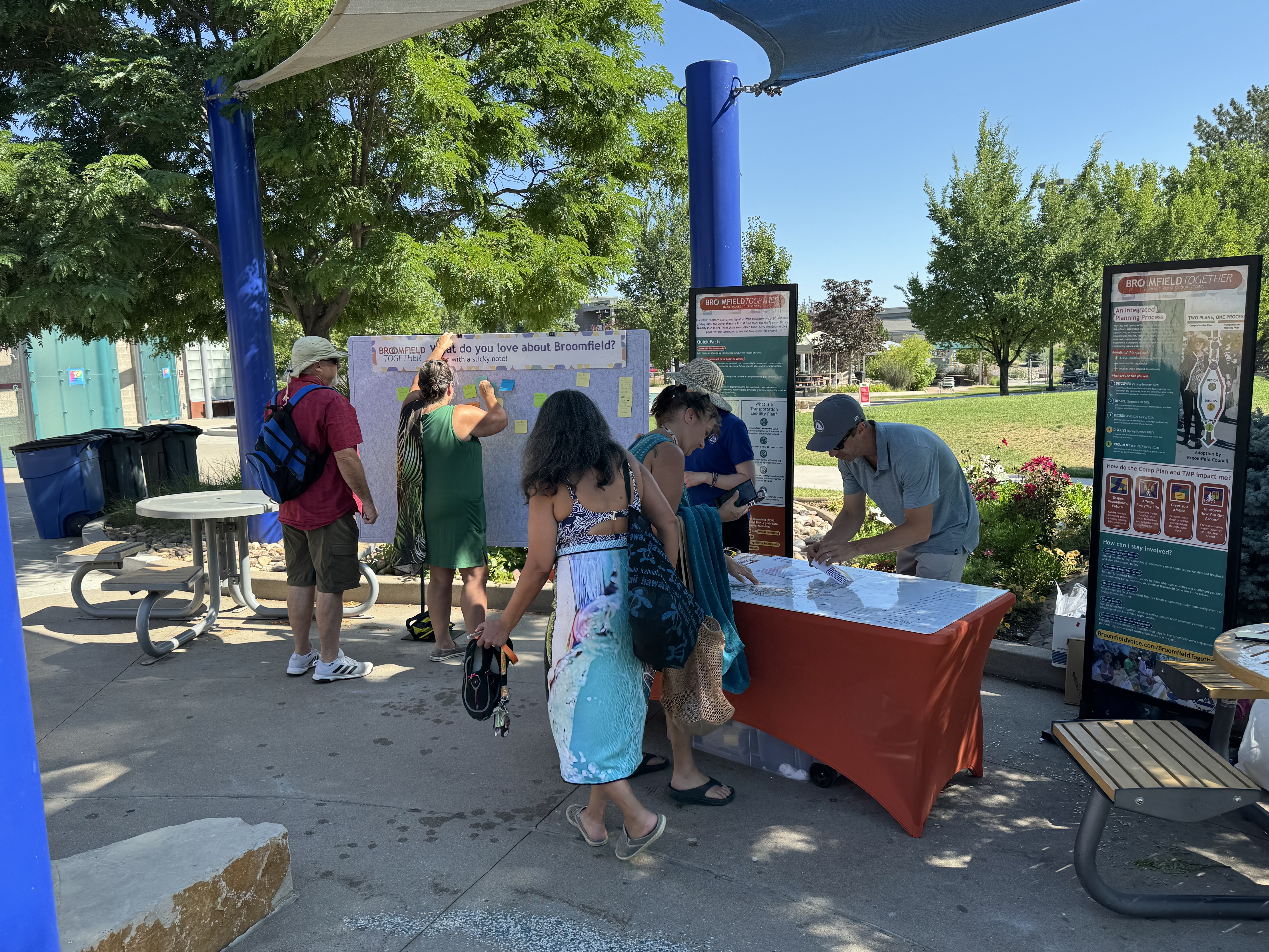 Photograph of an outdoor community engagement event for "Broomfield Together." People wearing swim suits and covers are interacting with a large gray board that asks, "What do you love about Broomfield? Tell us with a sticky note!"