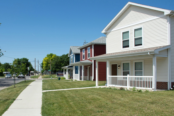Row of colorful houses facing a green lawn, sidewalk, and residential street with cars.
