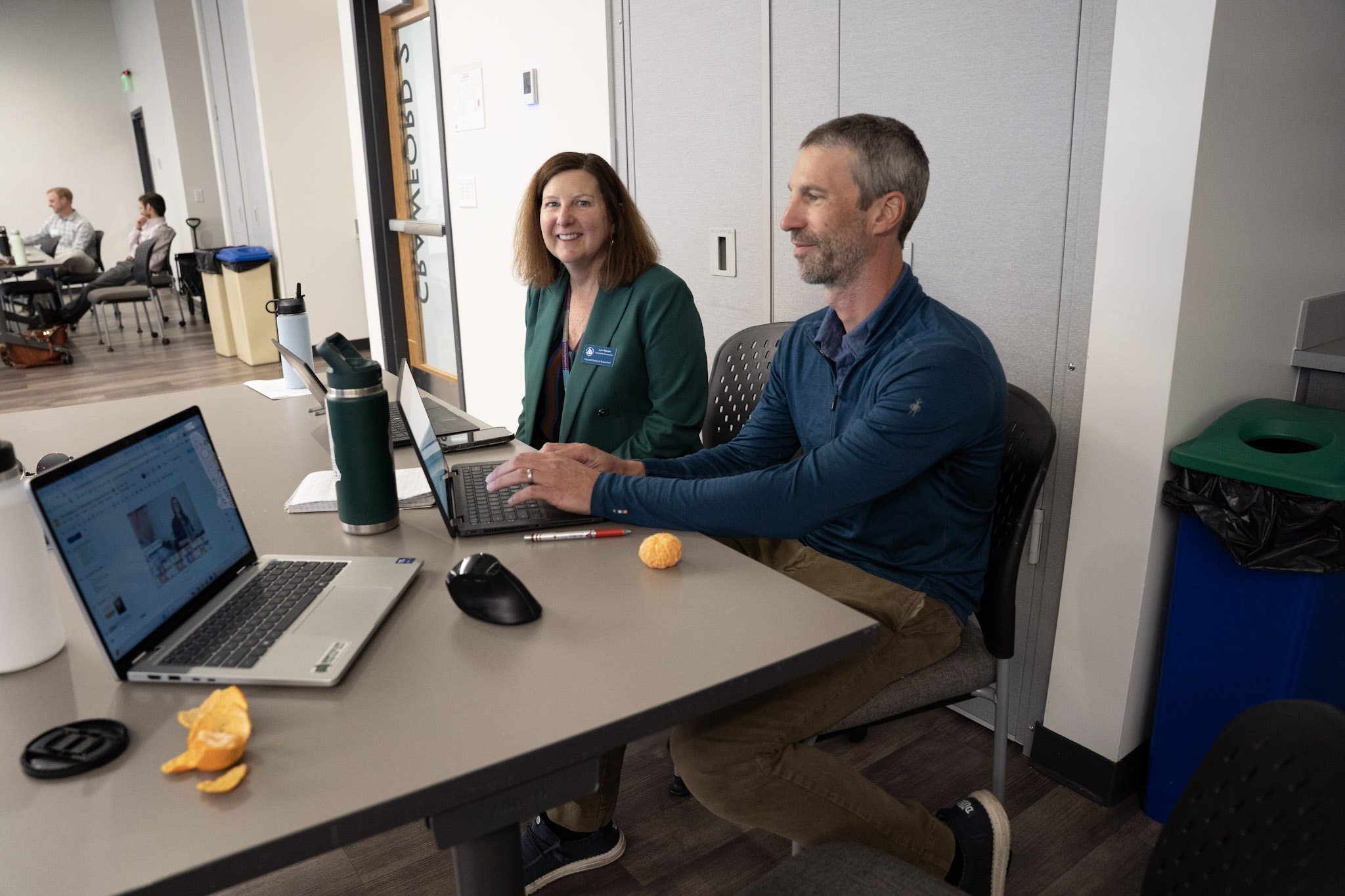 Two people sit working at a table. The woman is smiling at the camera while the man is typing on his lap top.