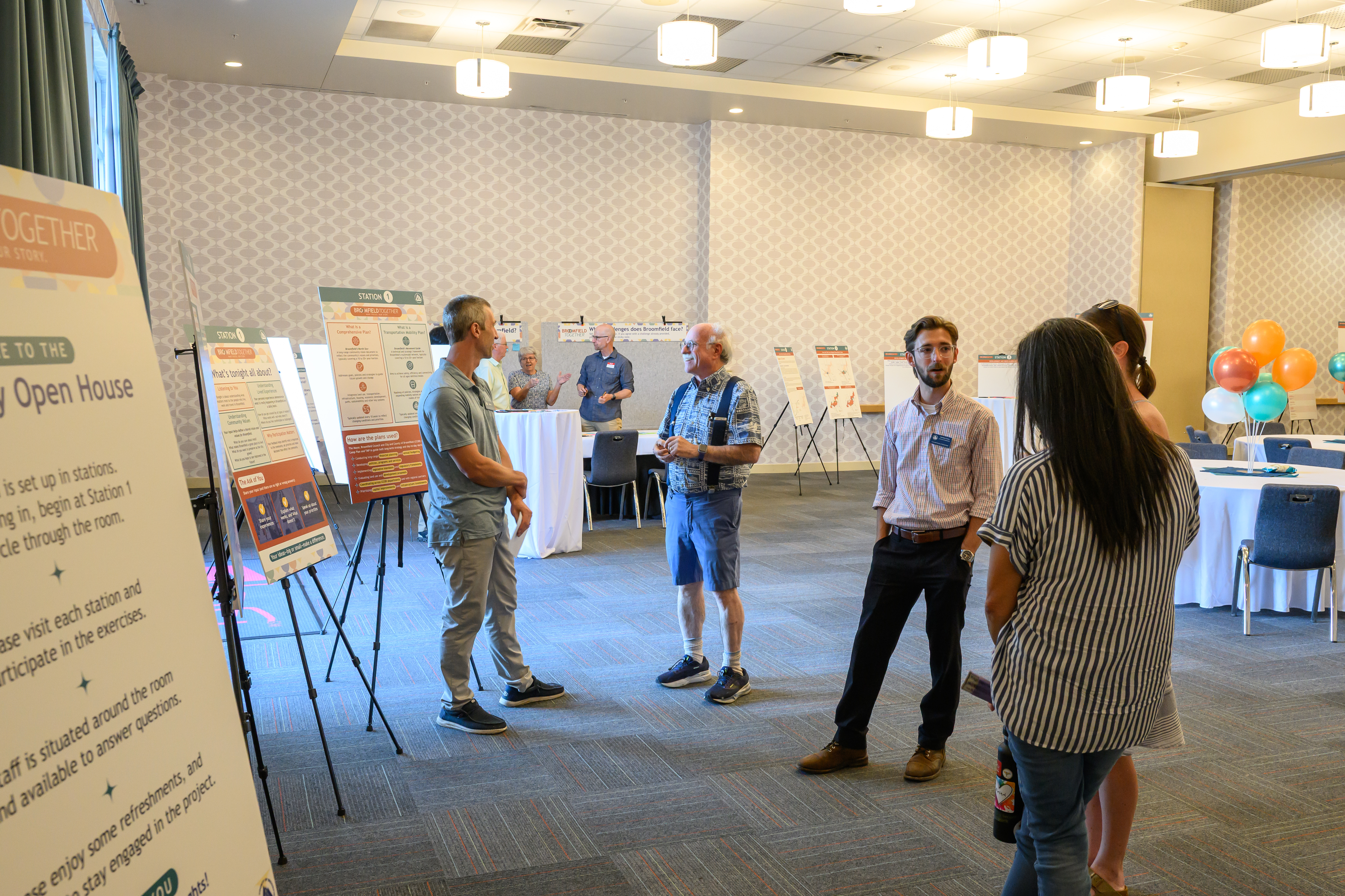 A photograph of a community open house event in a large, well-lit room. Several people are engaged in conversation near informational posters mounted on easels. A large sign in the foreground welcomes attendees, explaining that the event is organized into stations and encouraging participation.