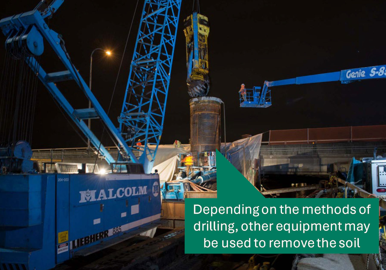 A drill with a scoop is suspended by a crane above a steel casing. 