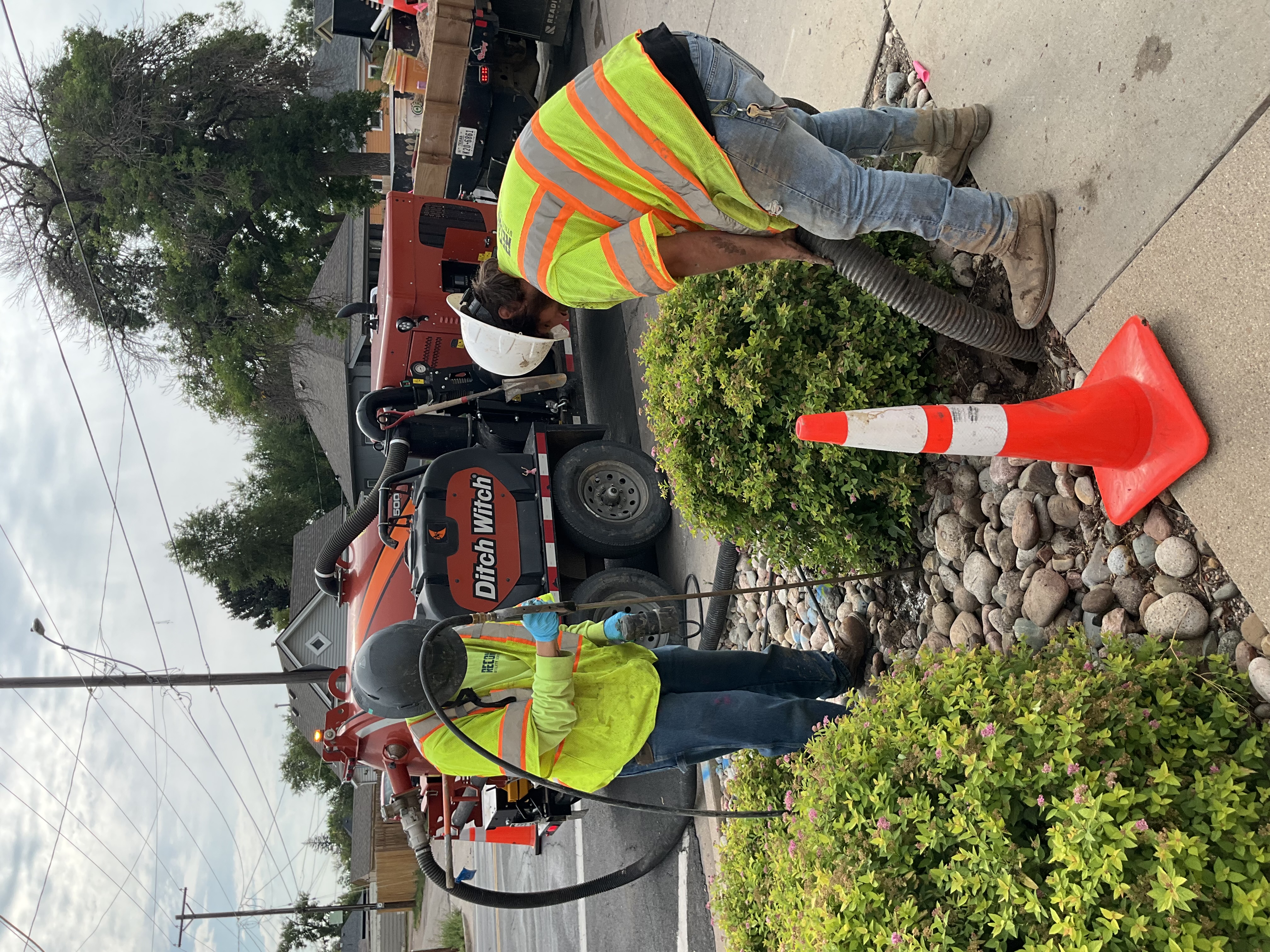 Two workers wearing safety vests and helmets use a high-water pressure and vacuum hose to create a hole to visually inspect a water service line.