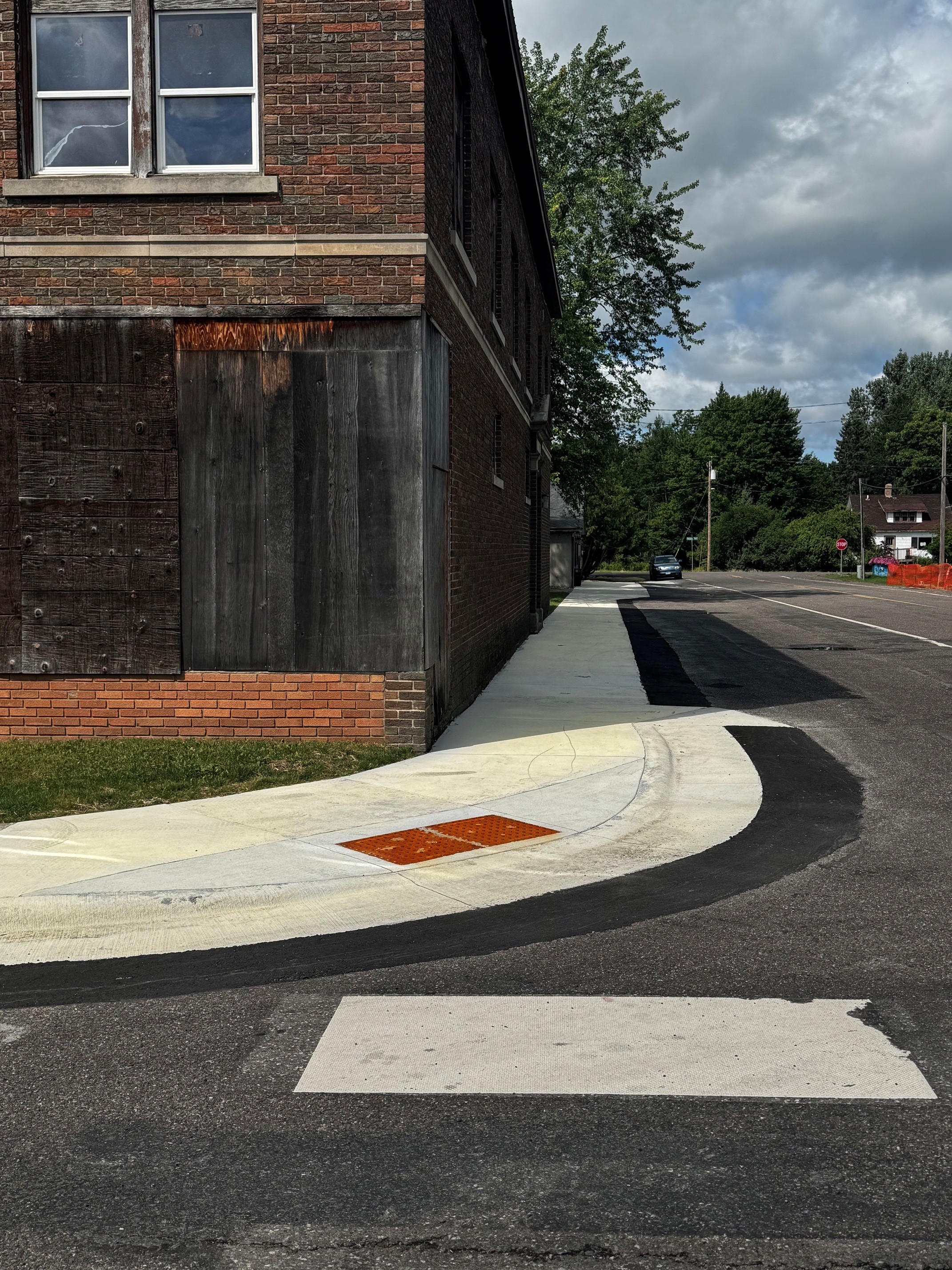 A weathered brick building with a sidewalk that curves around a corner, featuring a tactile orange pad. A blue car is parked on the street amid greenery.