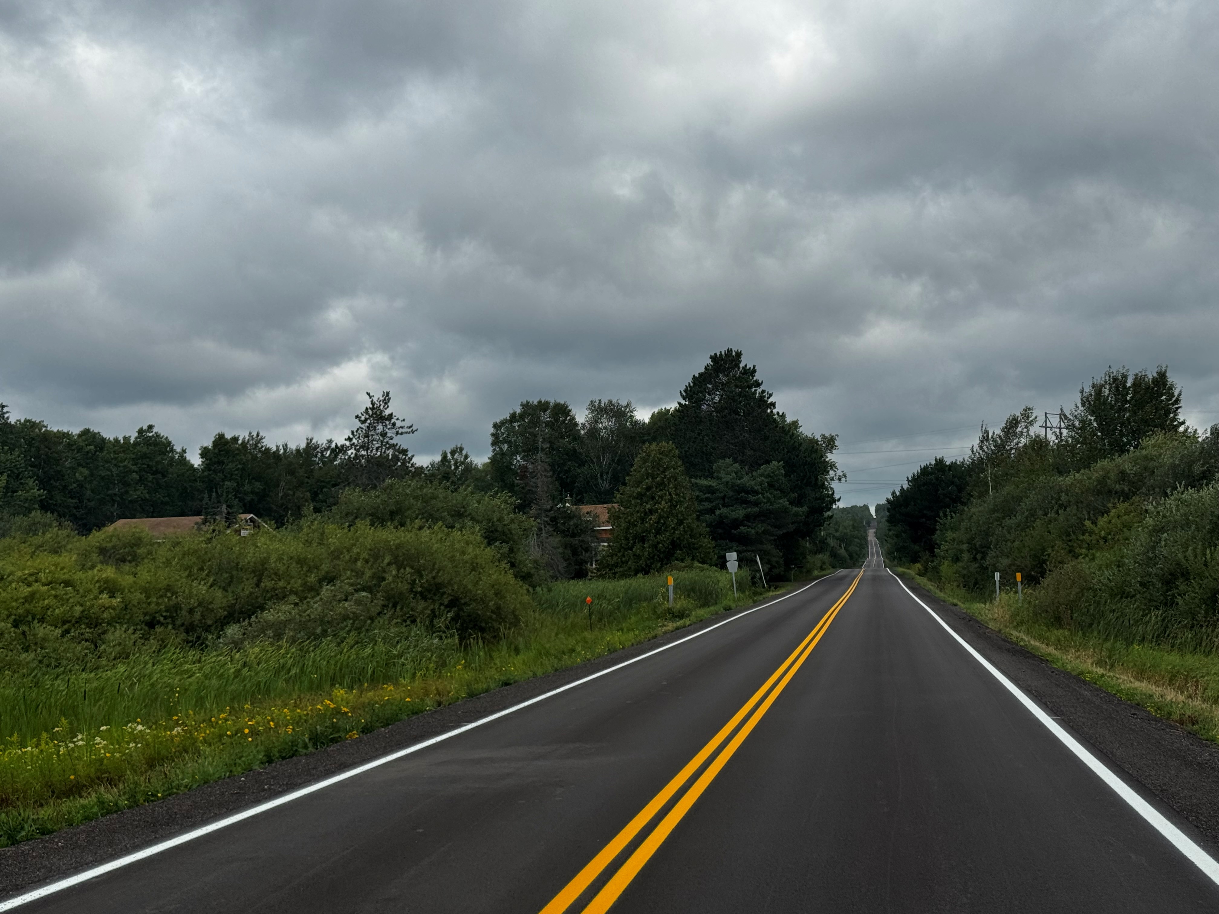 A freshly paved rural road with double yellow center lines leads through dense greenery beneath dark storm clouds.