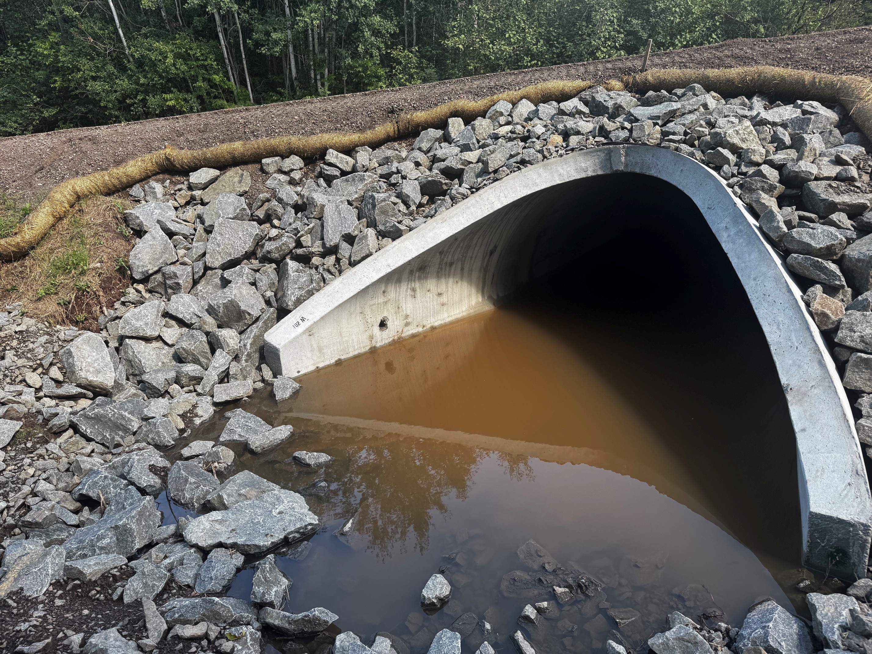 Concrete culvert surrounded by gray rocks, with murky water in the foreground and a wooded background.