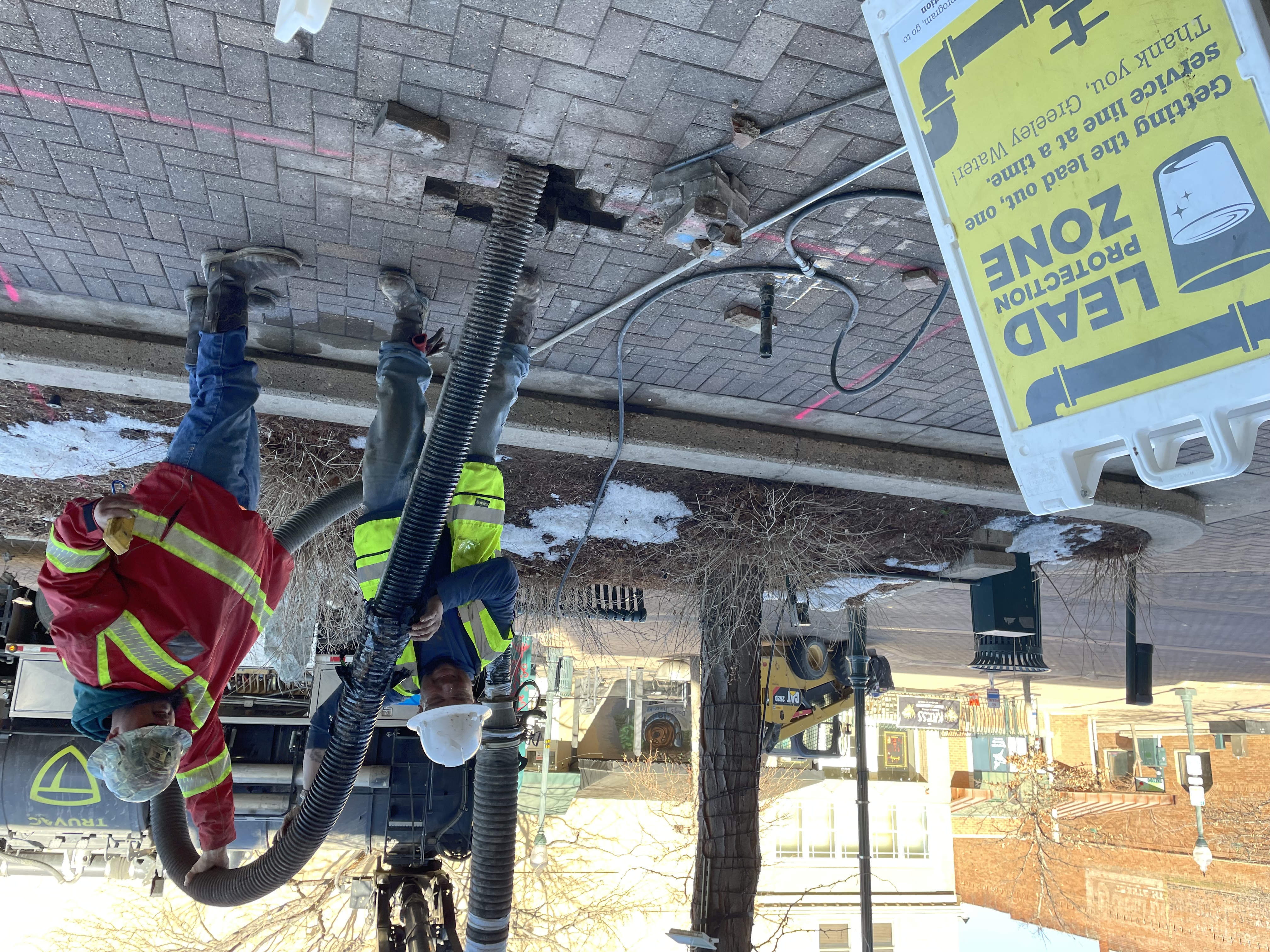 Two workers control a large vacuum hose to remove dirt from a hole through a brick-paved sidewalk.