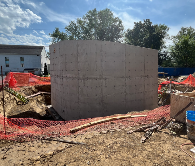 Wood footings in dirt from the last image are now filled with concrete and vertical walls have been poured and are standing tall. Construction fence and materials in the foreground.