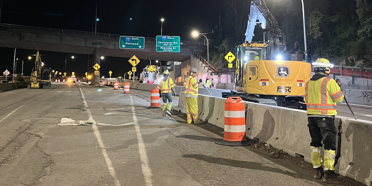 Nighttime road construction scene with workers, an excavator and traffic barrels near a highway overpass.