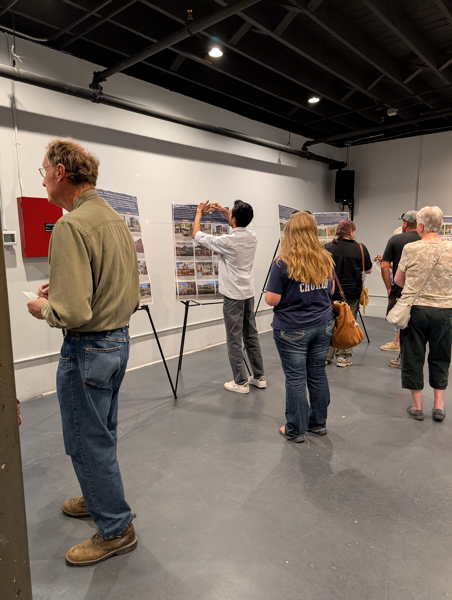 Six people stand in a large room looking at display boards on easels around the room. They are placing dots for the items they like or don't like.