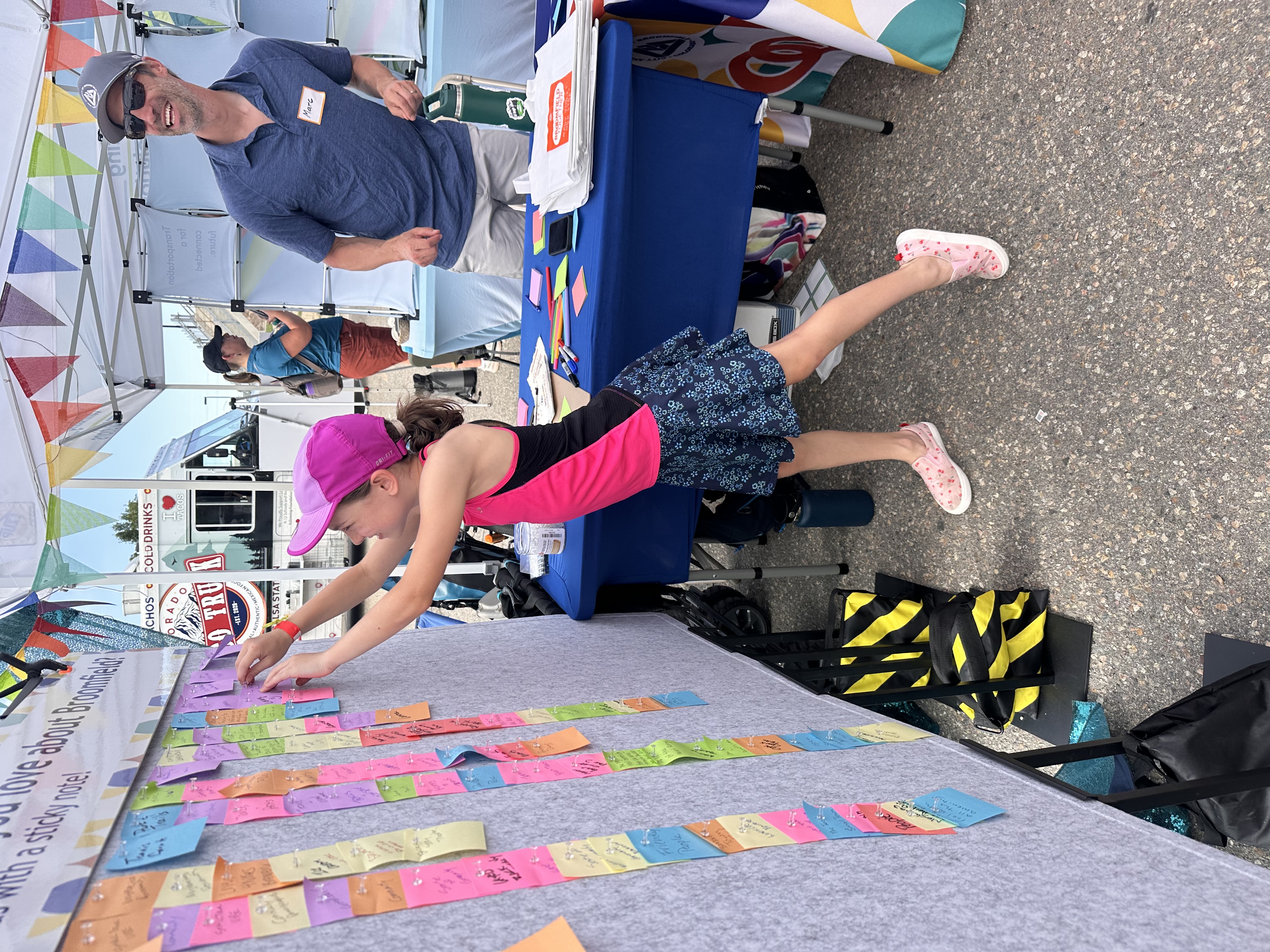 A young girl in a pink hat and tank top pins a sticky note to a large display board. The board is covered in numerous colorful handwritten notes. To the right, a man stands behind a table with event materials.