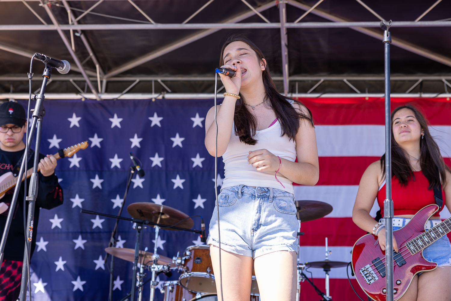 women in white tank top and denim shorts singing into a mic with 2 guitarists and an American flag behind her.