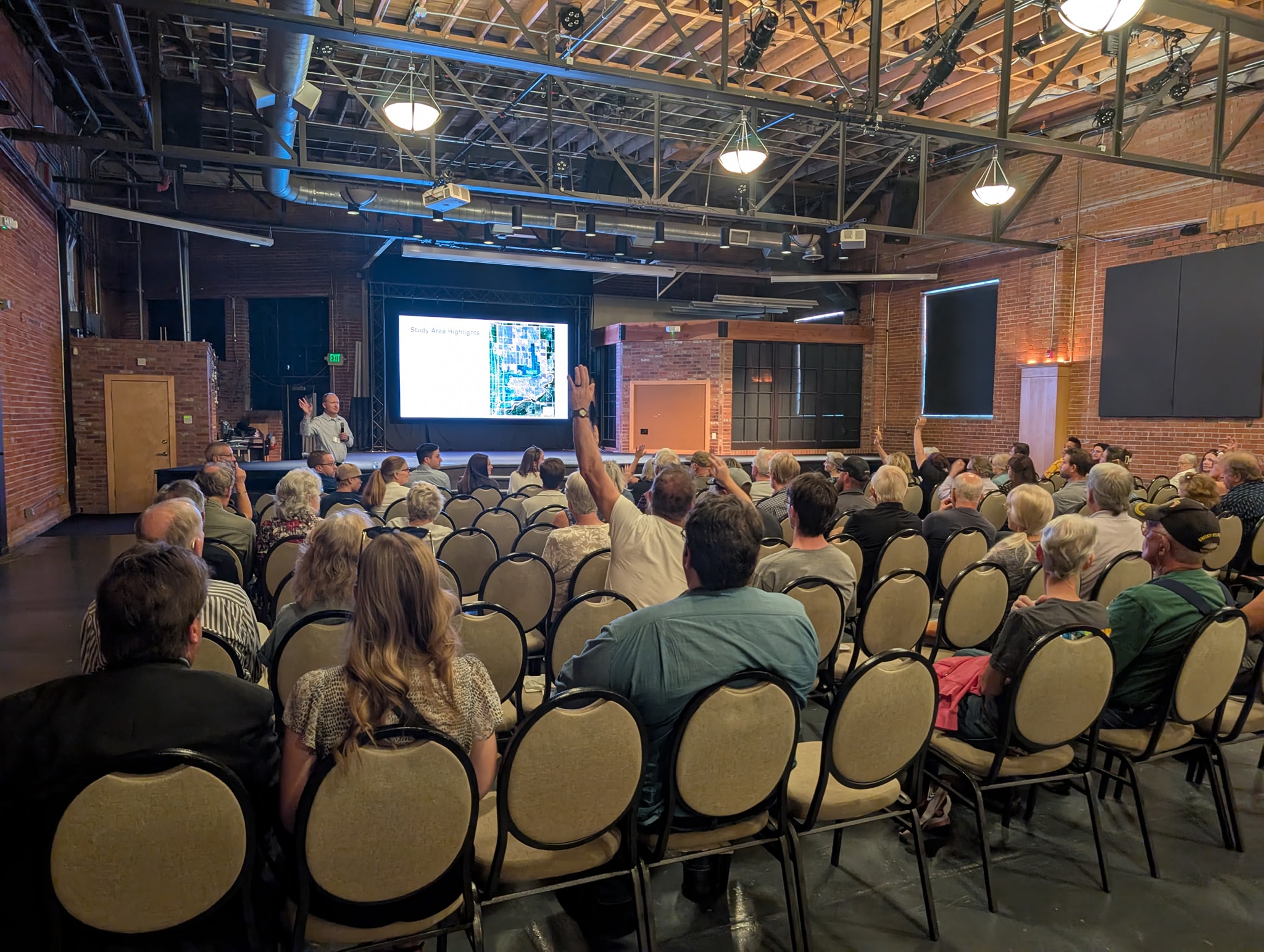 A large crowd seated in rows listenting to a presentation. There is a large screen on a stage. Some people have their hands raised.