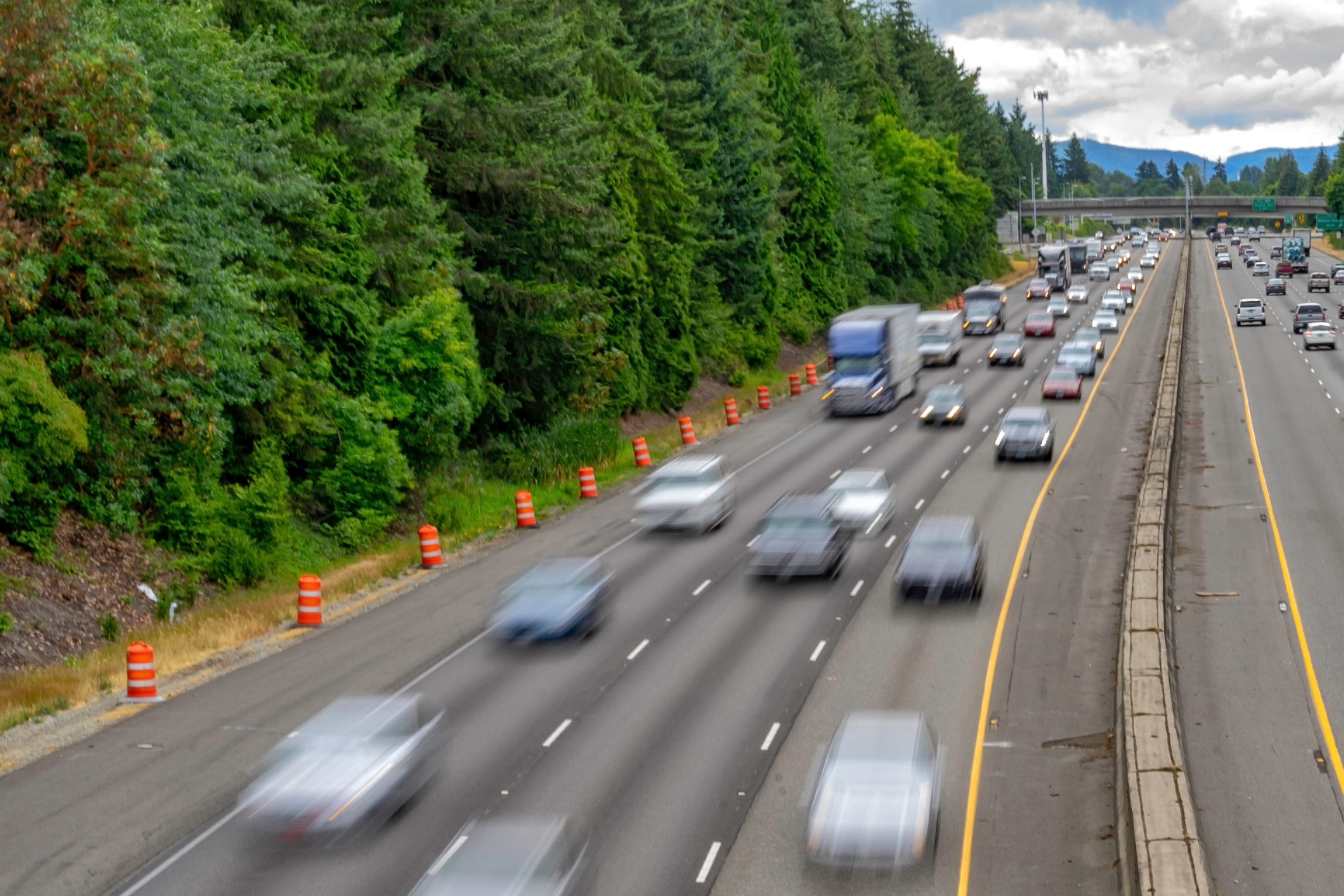An elevated view of Interstate 5 in Washington, with motion-blurred traffic moving in both directions, a semi-truck among the cars, orange construction barrels lining the forested shoulder, and an overpass and hills in the distance.