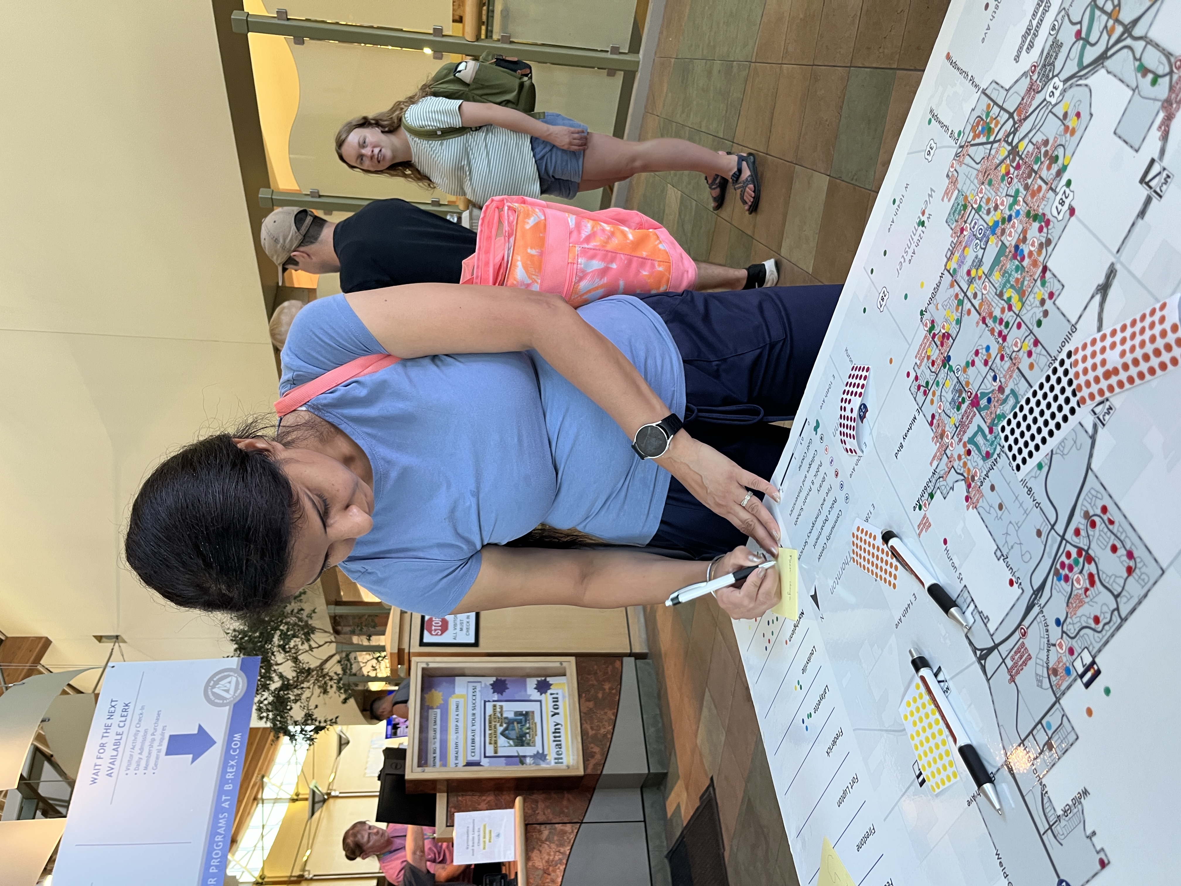 A woman in a blue t-shirt writes on a yellow sticky note placed on a large street map spread across a table. The map is marked with many small, colored dot stickers, indicating a community feedback activity.