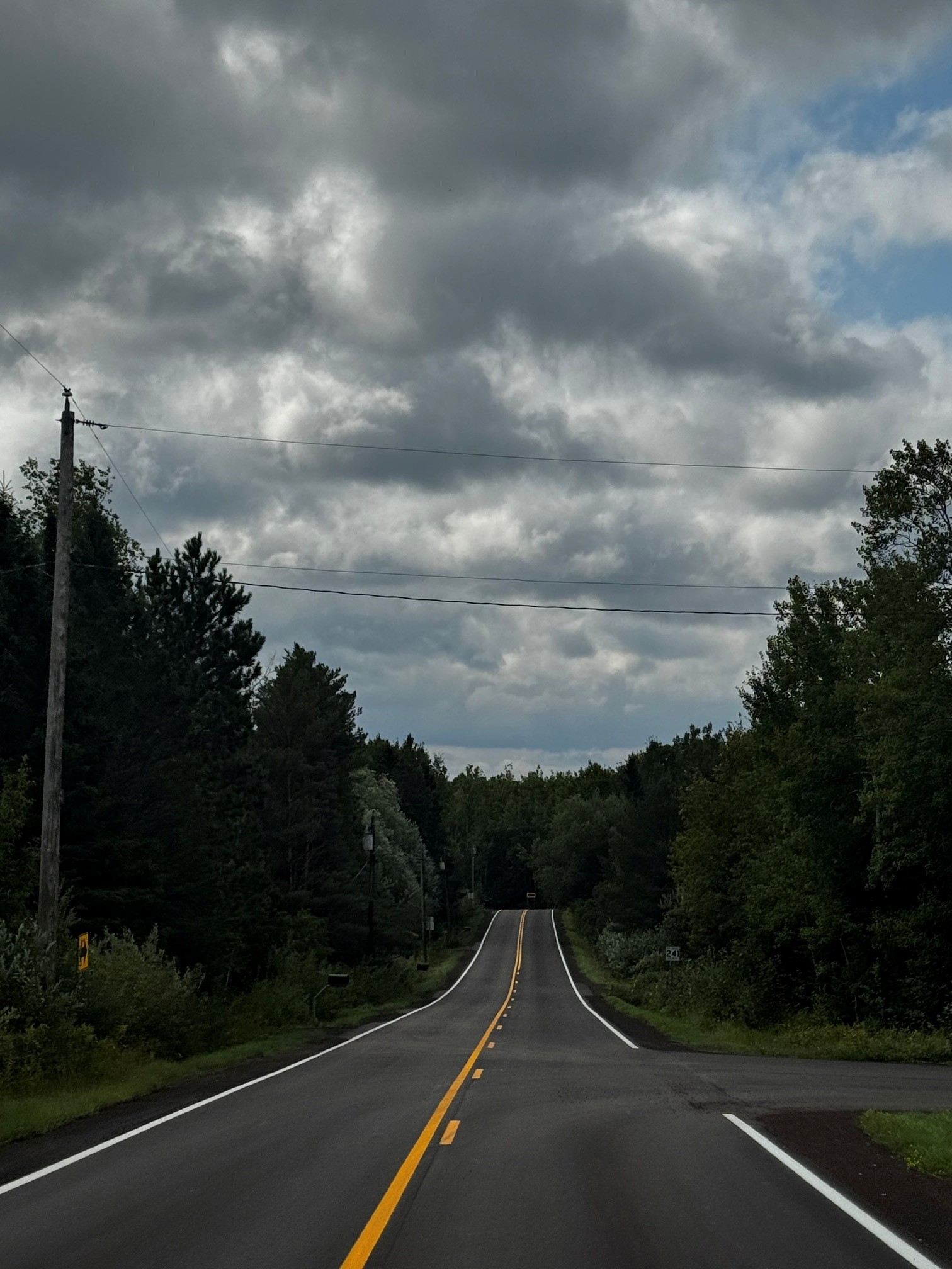 A tree-lined two-lane road leads toward dense woods beneath dark, layered storm clouds, with utility wires crossing the sky.