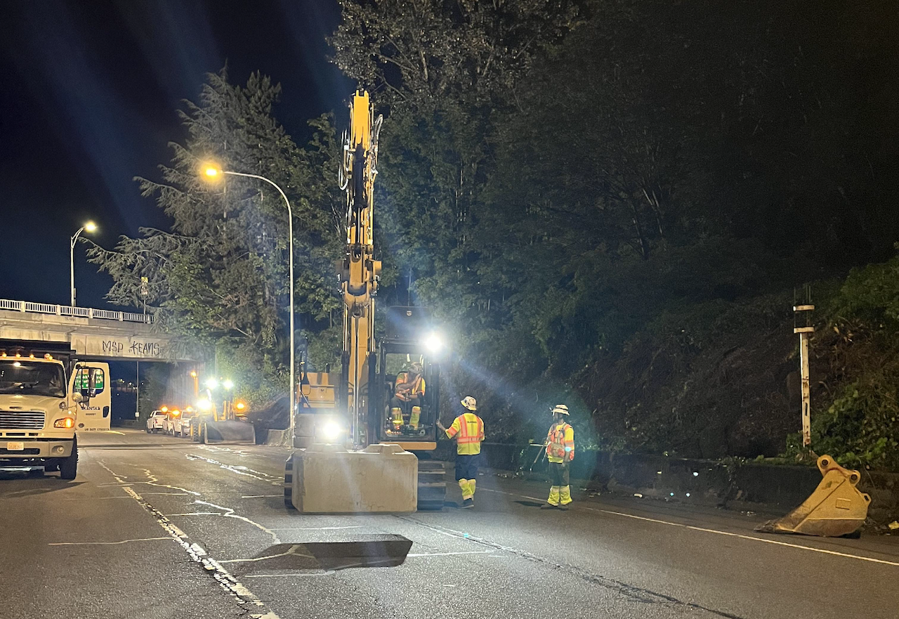 Nighttime road construction scene with workers, a truck and machinery moving a concrete traffic block.