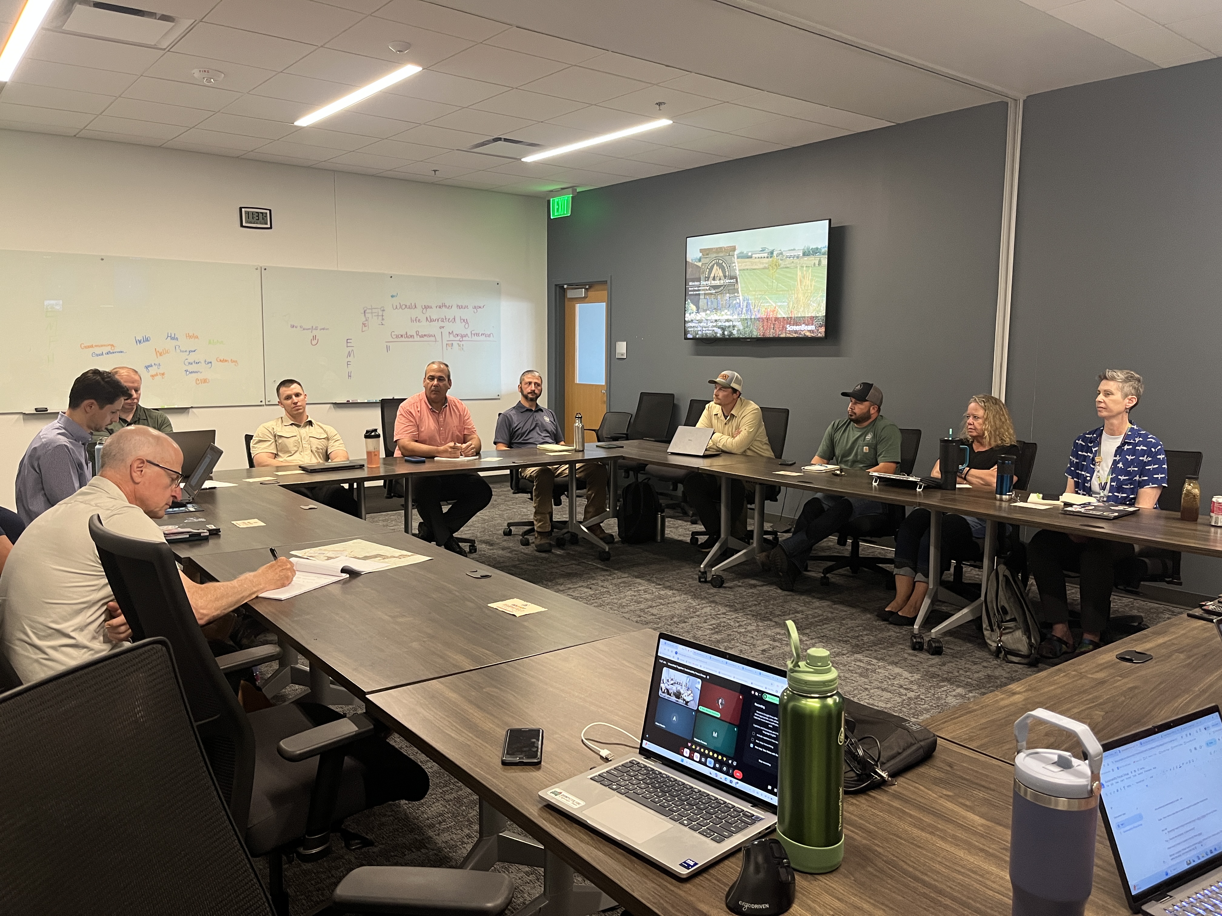 Photograph of a professional meeting taking place in a conference room with people seated around a large U-shaped table. In the foreground, a person writes on a notepad. Other attendees, dressed in business-casual attire, listen and look toward a wall-mounted monitor showing a presentation slide. The background features whiteboards with handwritten notes. Several laptops, notebooks, and water bottles are scattered across the table.