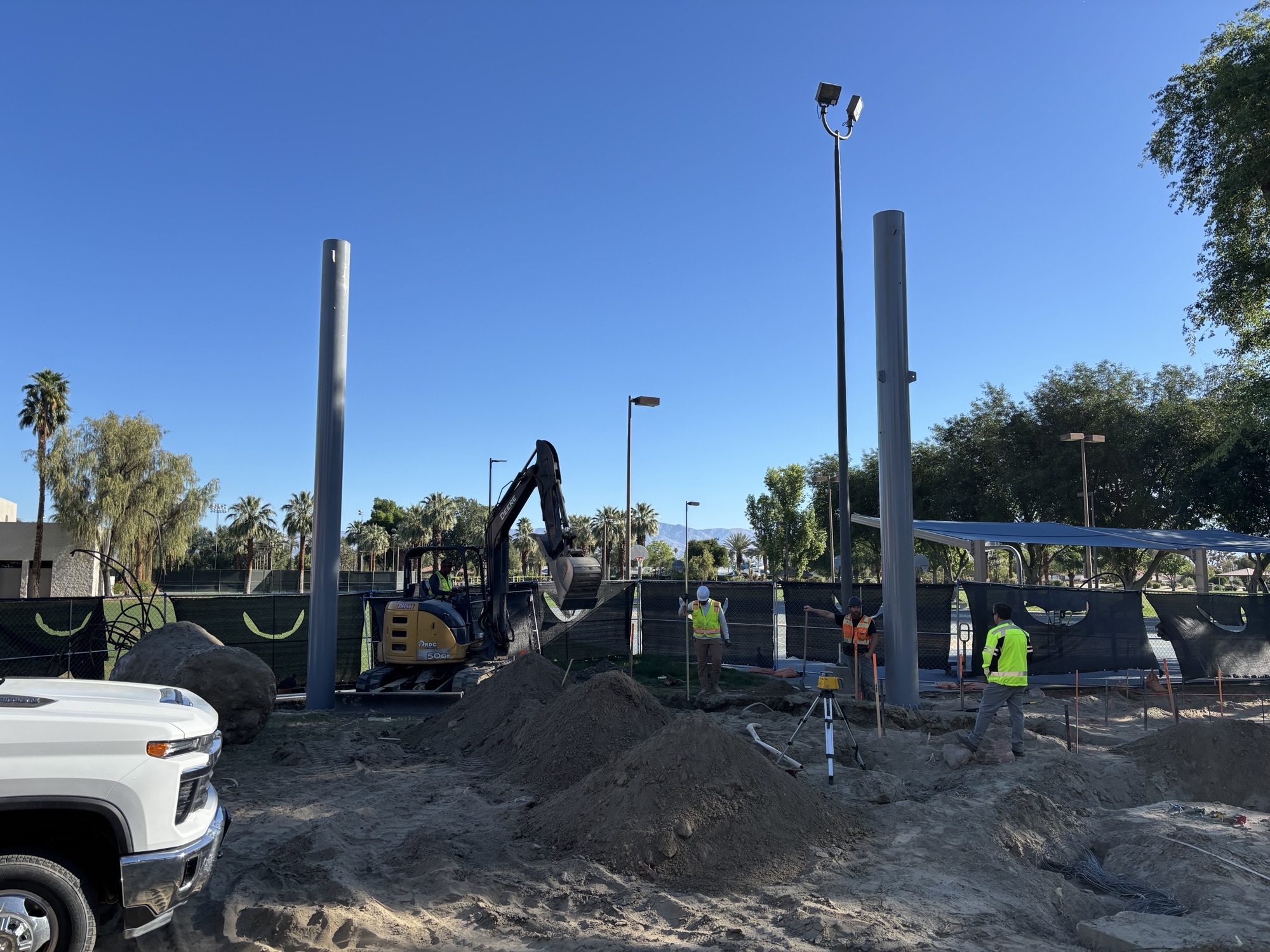 Workers in safety vests operate a small excavator and prepare footings around tall steel columns at an outdoor construction site near a basketball court.