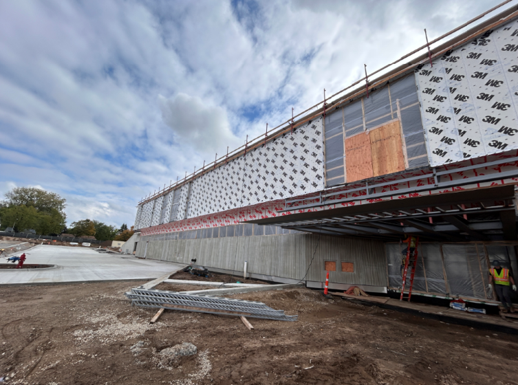 Image shows building under construction on the left side of the image, materials include concrete, metal, and eventually glass. Approach to building coming from the left side of the image is dirt awaiting sidewalks and landscaping.