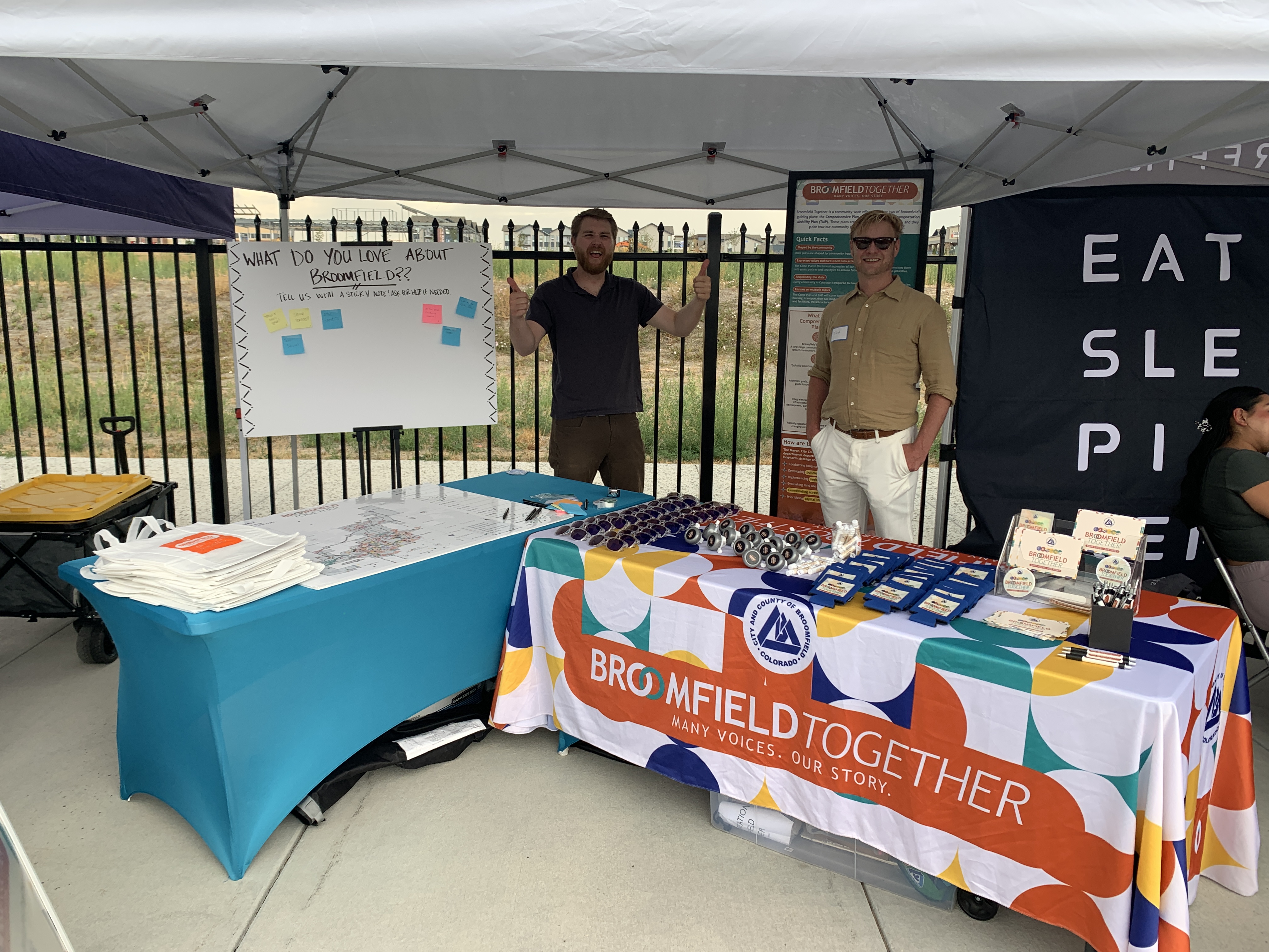 Photograph of two men at a "Broomfield Together" community outreach booth under a white tent. On the left, a man gives two thumbs up next to a whiteboard that says, "WHAT DO YOU LOVE ABOUT BROOMFIELD?? TELL US WITH A STICKY NOTE!" On the right, a man in sunglasses stands behind a table covered with a colorful tablecloth reading "BROOMFIELD TOGETHER MANY VOICES. OUR STORY." and featuring the CCOB logo. The tables are stocked with maps, brochures, and promotional items.