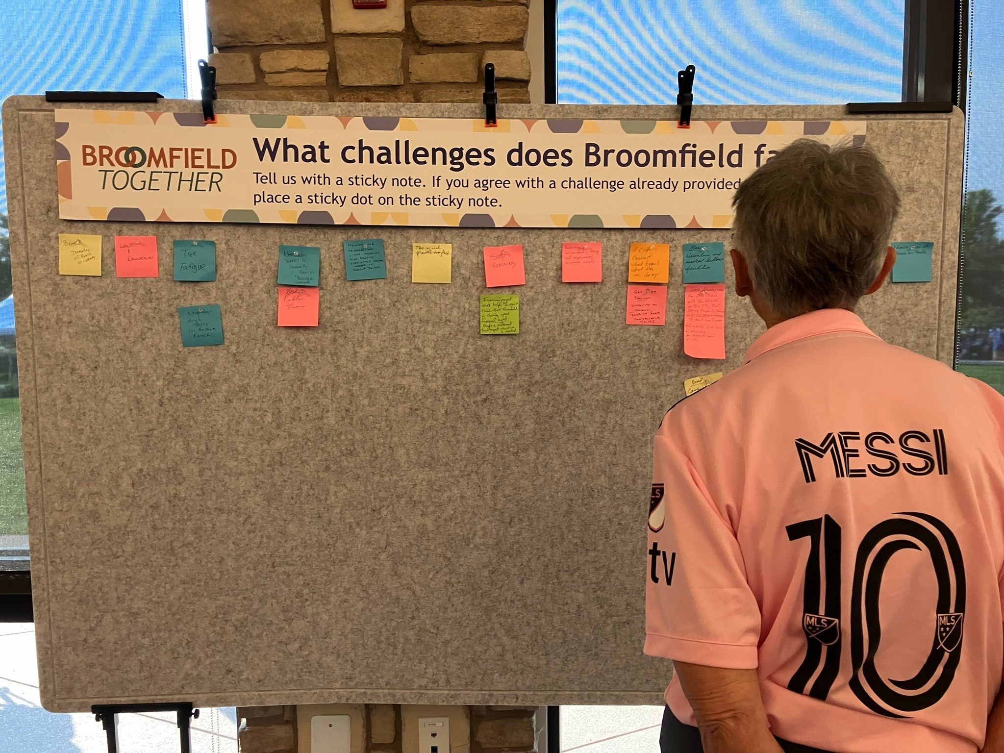 Photograph of a person from behind, wearing a pink Lionel Messi #10 jersey, looking at a community feedback board. A banner at the top of the board reads "BROOMFIELD TOGETHER" and asks, "What challenges does Broomfield face?"