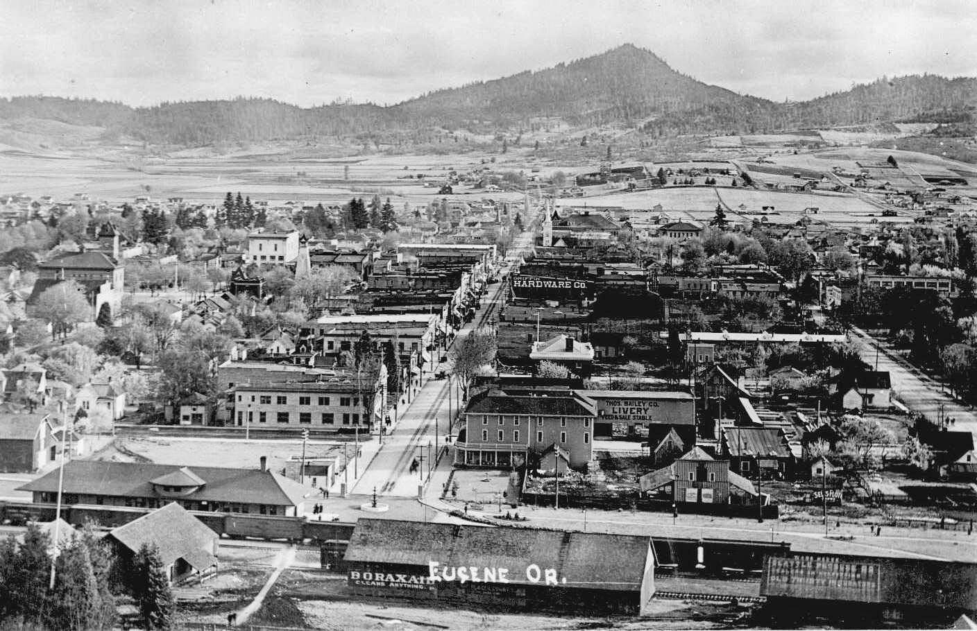 A black and white historic photo of Eugene taken from Skinner's Butte looking south down Willamette Street. It shows old buildings, farms, and mountains.