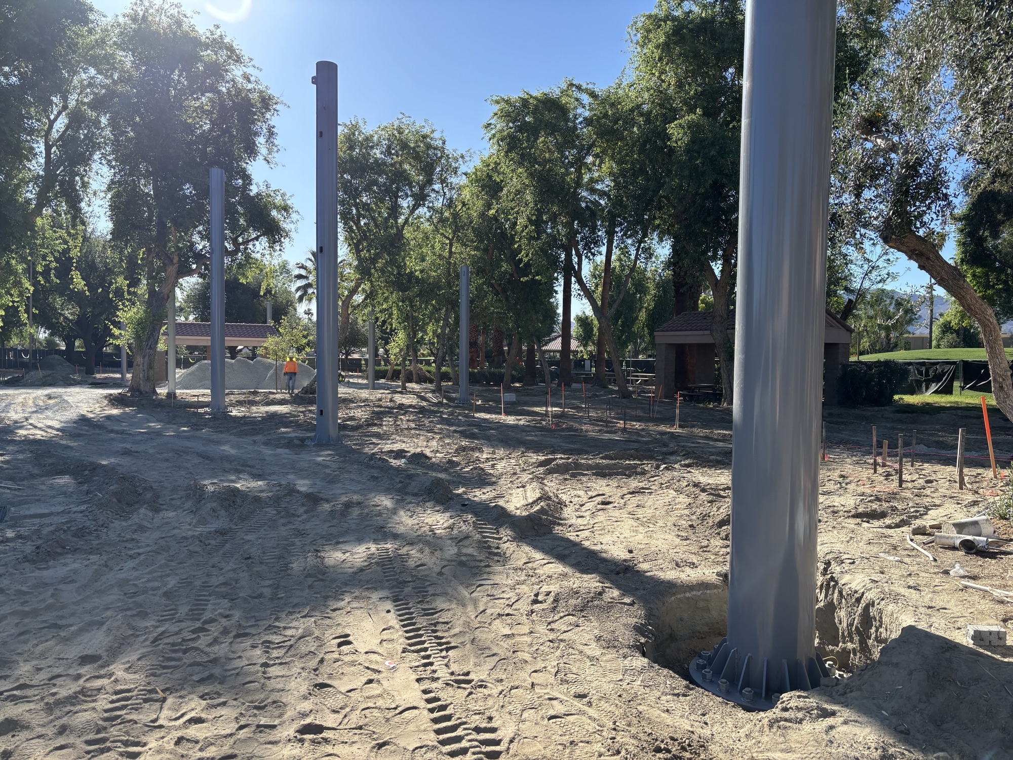 Construction site in a park with multiple tall steel columns installed in sandy ground, surrounded by trees and a small pavilion in the background.