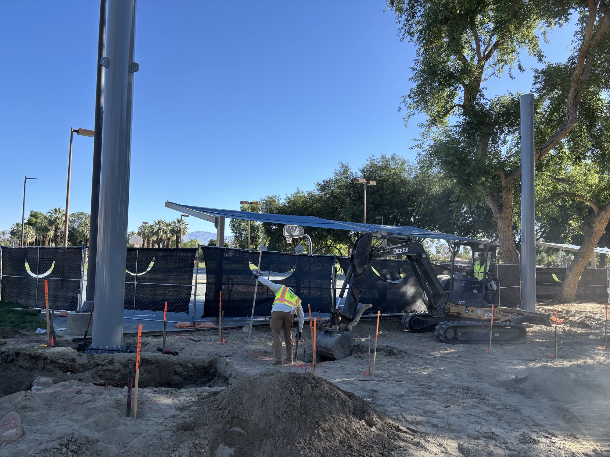 Crew members dig trenches and mark layout points beside installed steel columns, with a shade structure and fencing visible behind them.
